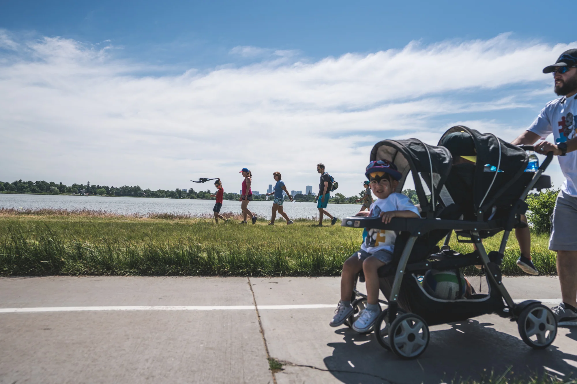Group of people walking around Sloan's Lake as a little boy swings a jacket above his head and the Denver skyline looms in the background