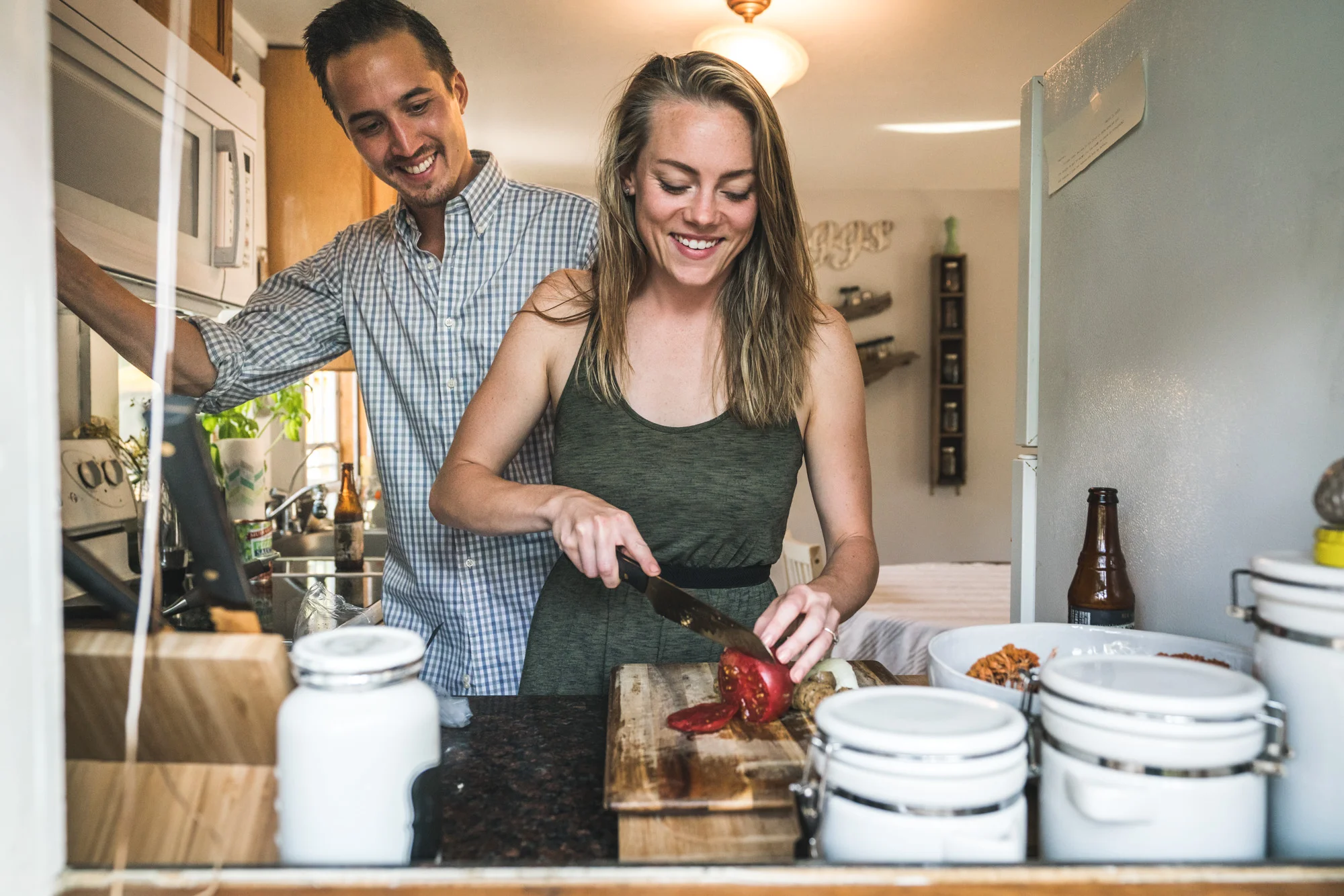 At home engagement session photo looking in a window at a man and woman smiling and cooking dinner together
