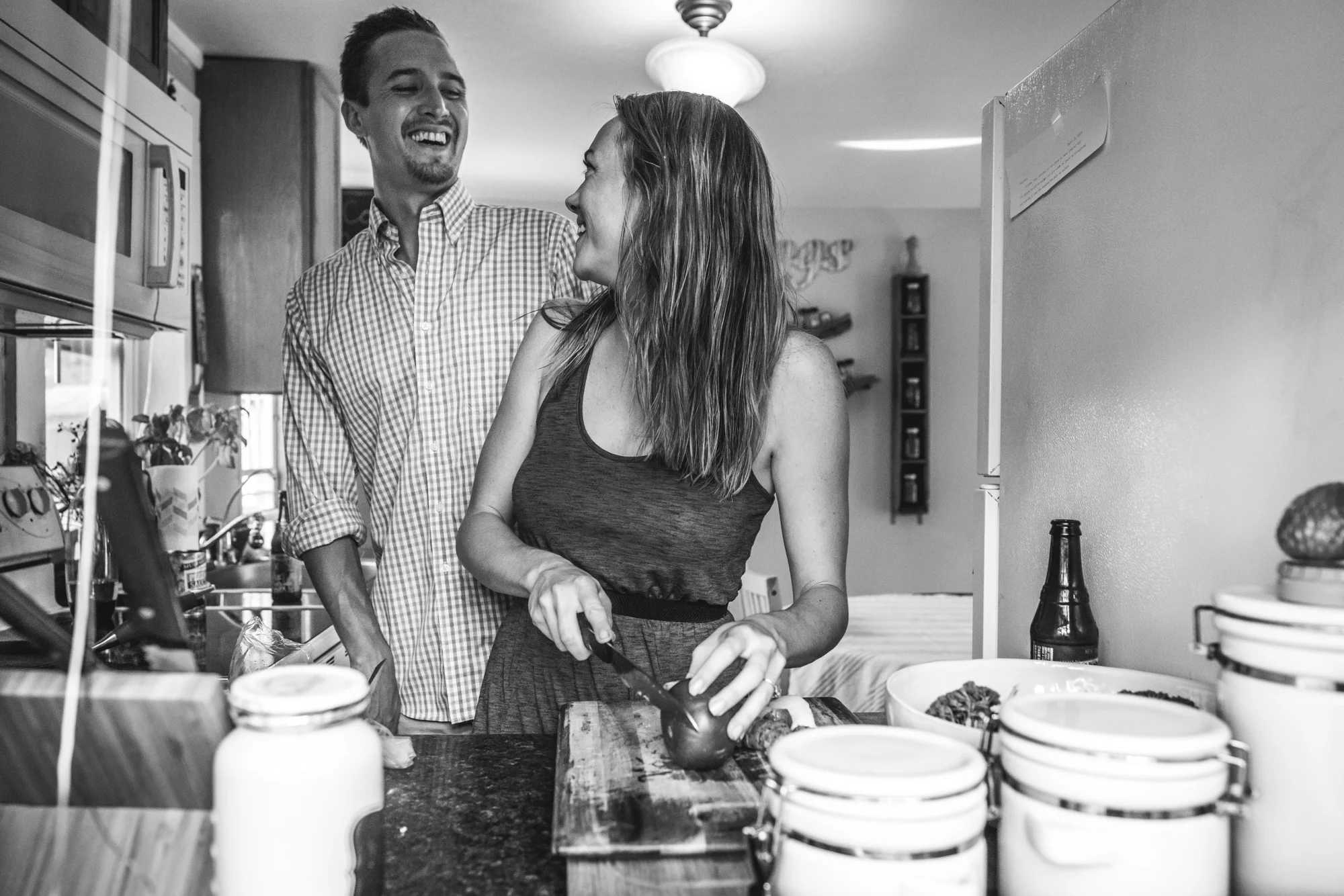 Black and white documentary engagement session photo looking in through a window at a man and woman coking dinner together and looking at each other and smiling