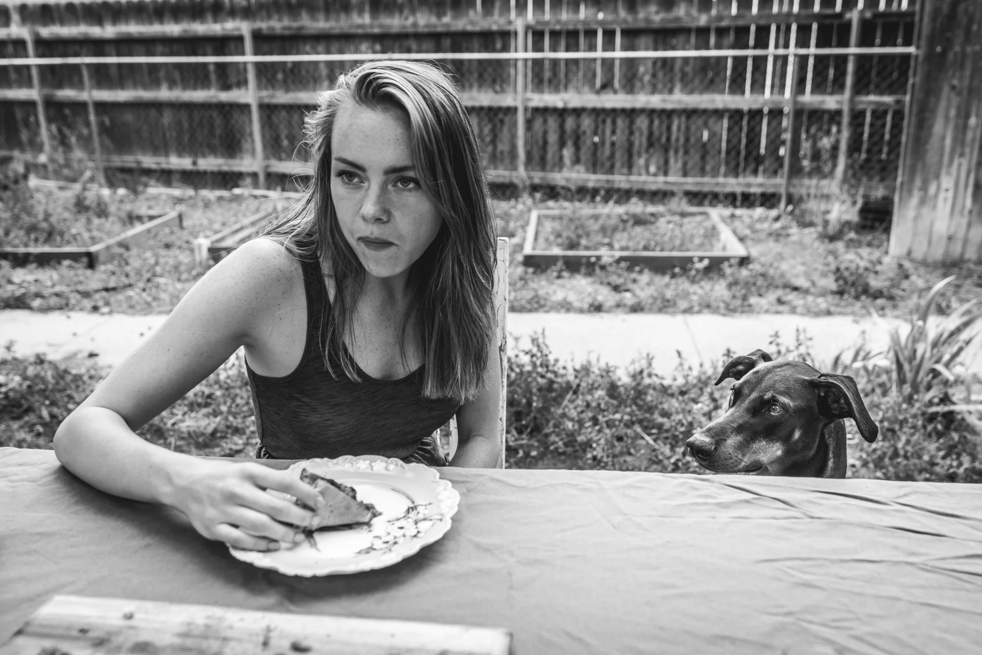 Black and white photo of a woman eating pizza at an outside table as her doberman pincher looks at the pizza longingly