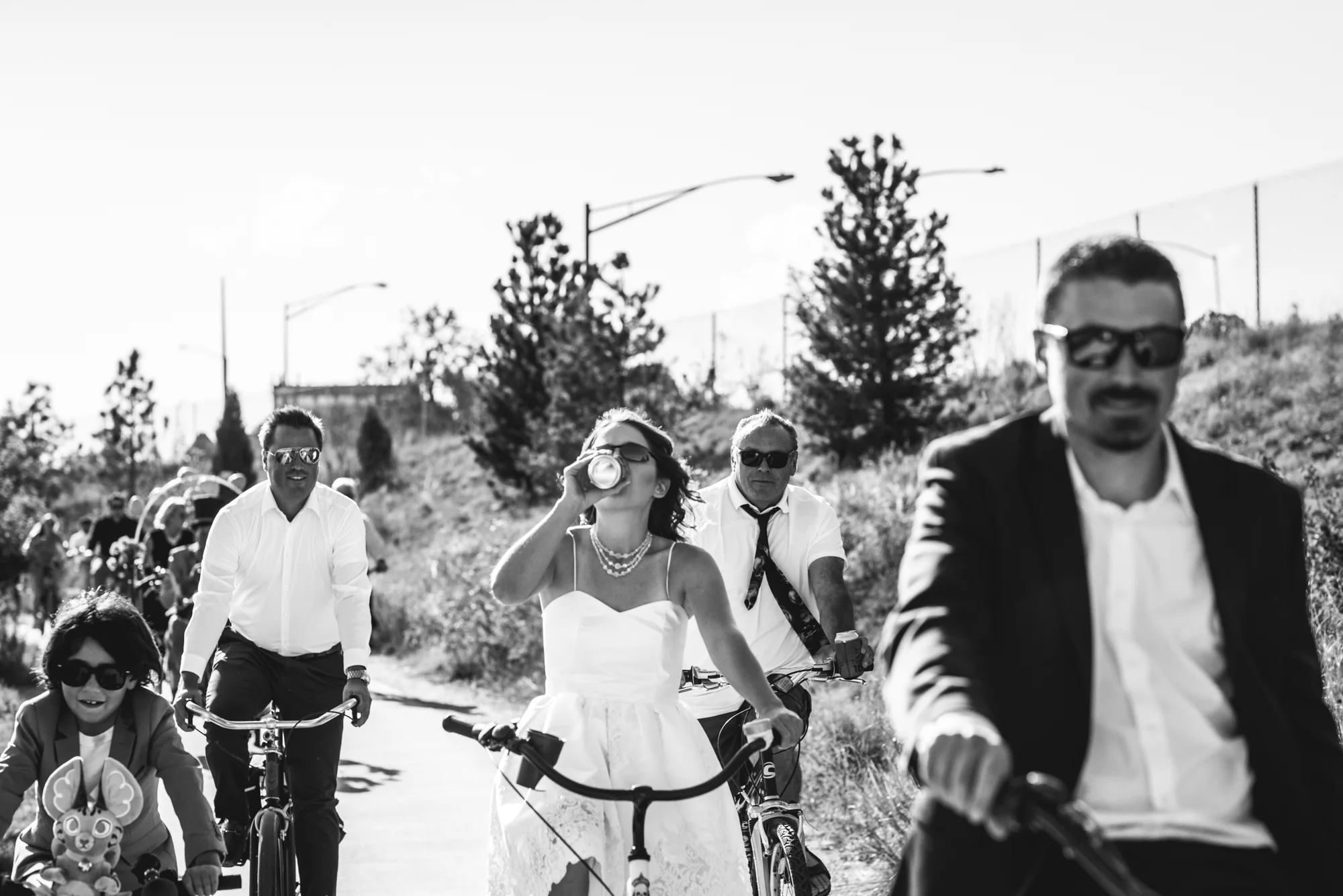 Black and white photo of a bride, groom, and their guests riding on bikes, the bride taking a swig of her beer