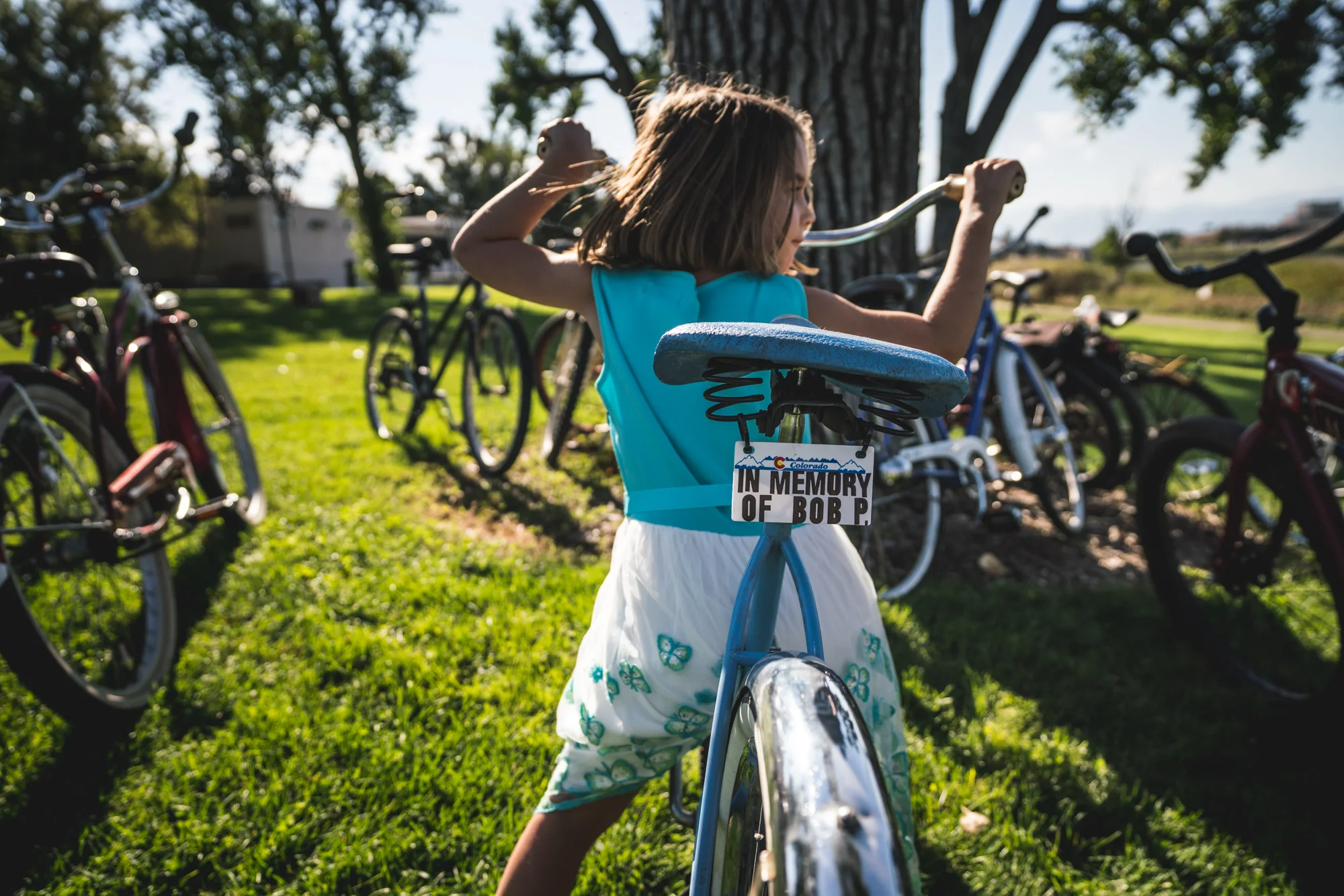 Little girl sits on a bike, surrounded by other bikes. The back of her bike bears a license plate that reads “In Memory of Bob P.” The little girl is wearing a fancy dress.