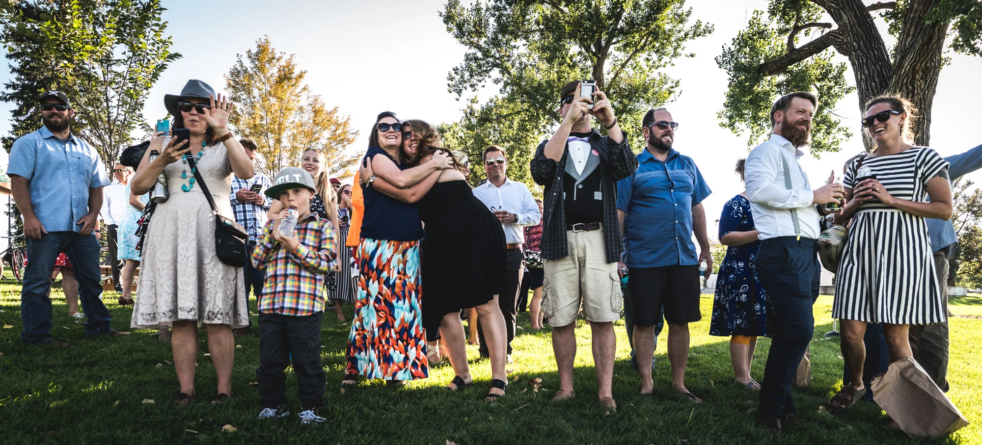 Wide shot of a bunch of wedding guests outdoors amongst trees smiling, hugging, and laughing