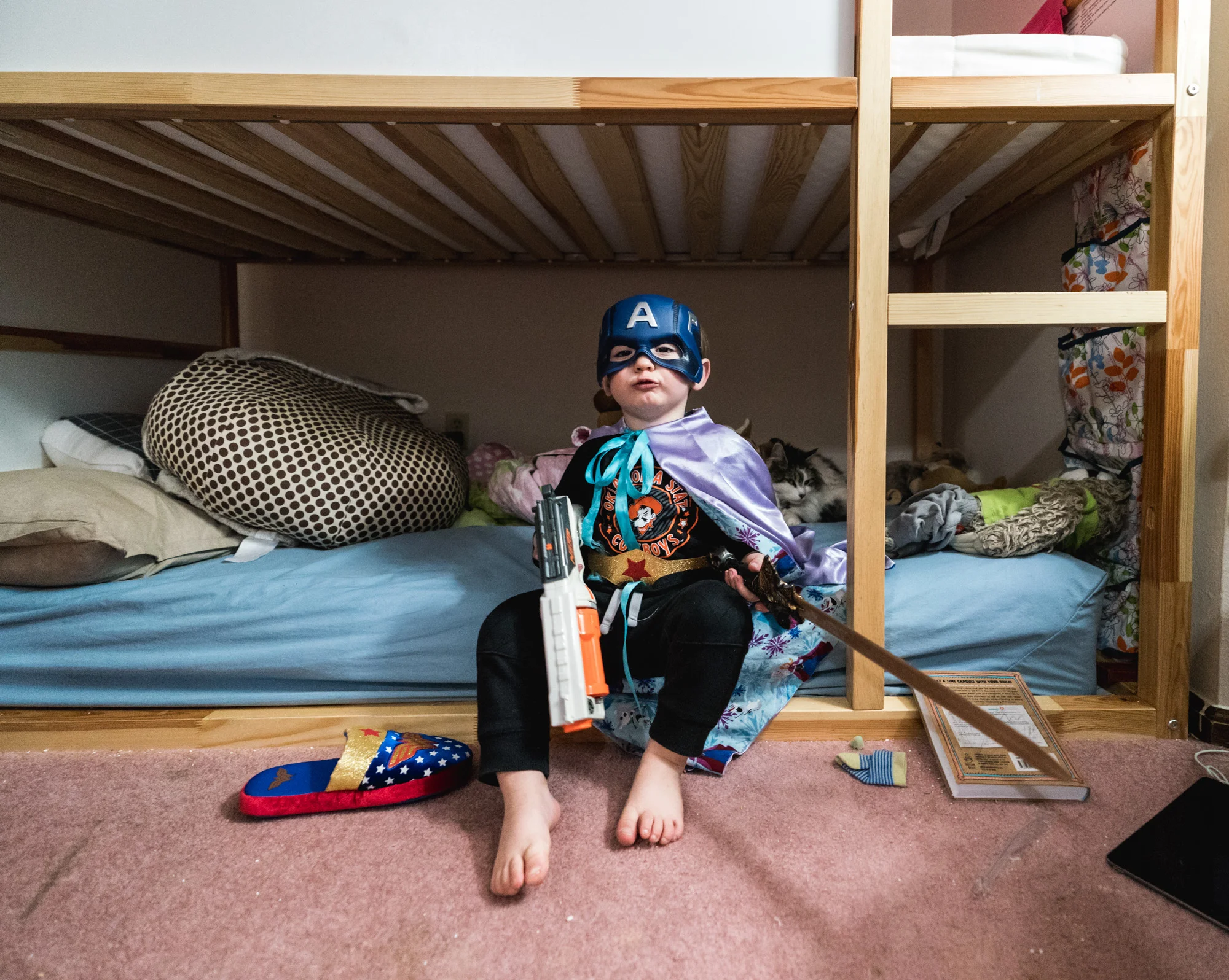 Little boy sitting on his bed wearing a superhero mask and cape and holding both a sword and a toy gun, looks straight to camera.