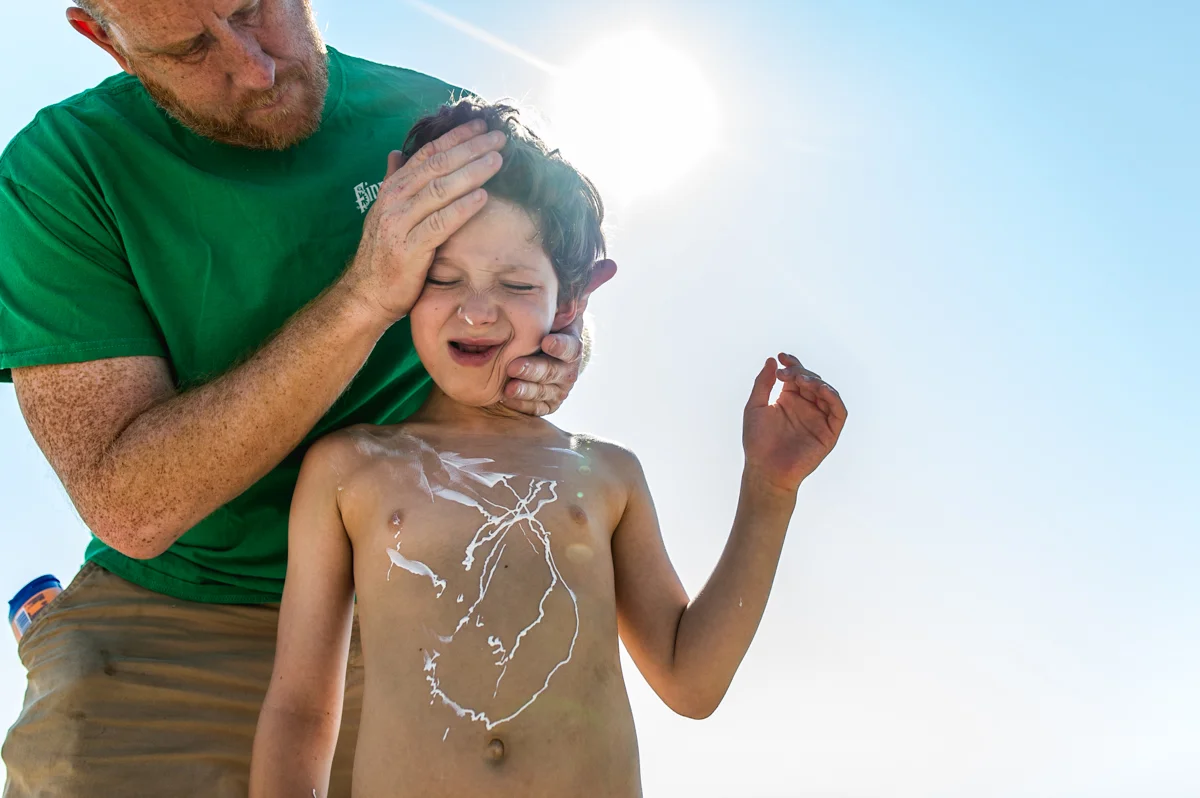 Little boy looks unhappy as his dad smears sunscreen all over his face