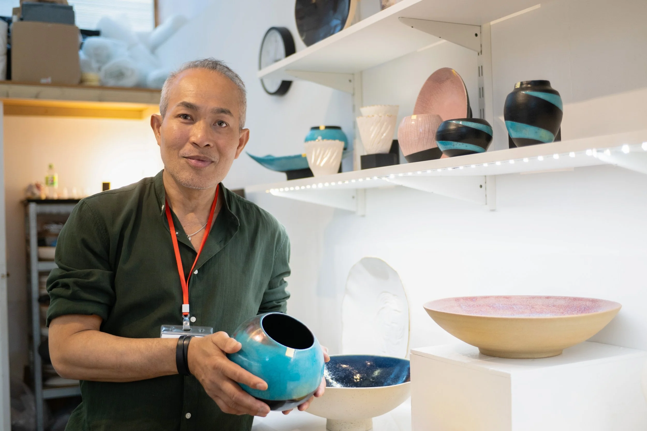 An older man with gray hair holding a blue and black ceramic bowl in a ceramics studio, surrounded by various finished ceramic pottery pieces on shelves.