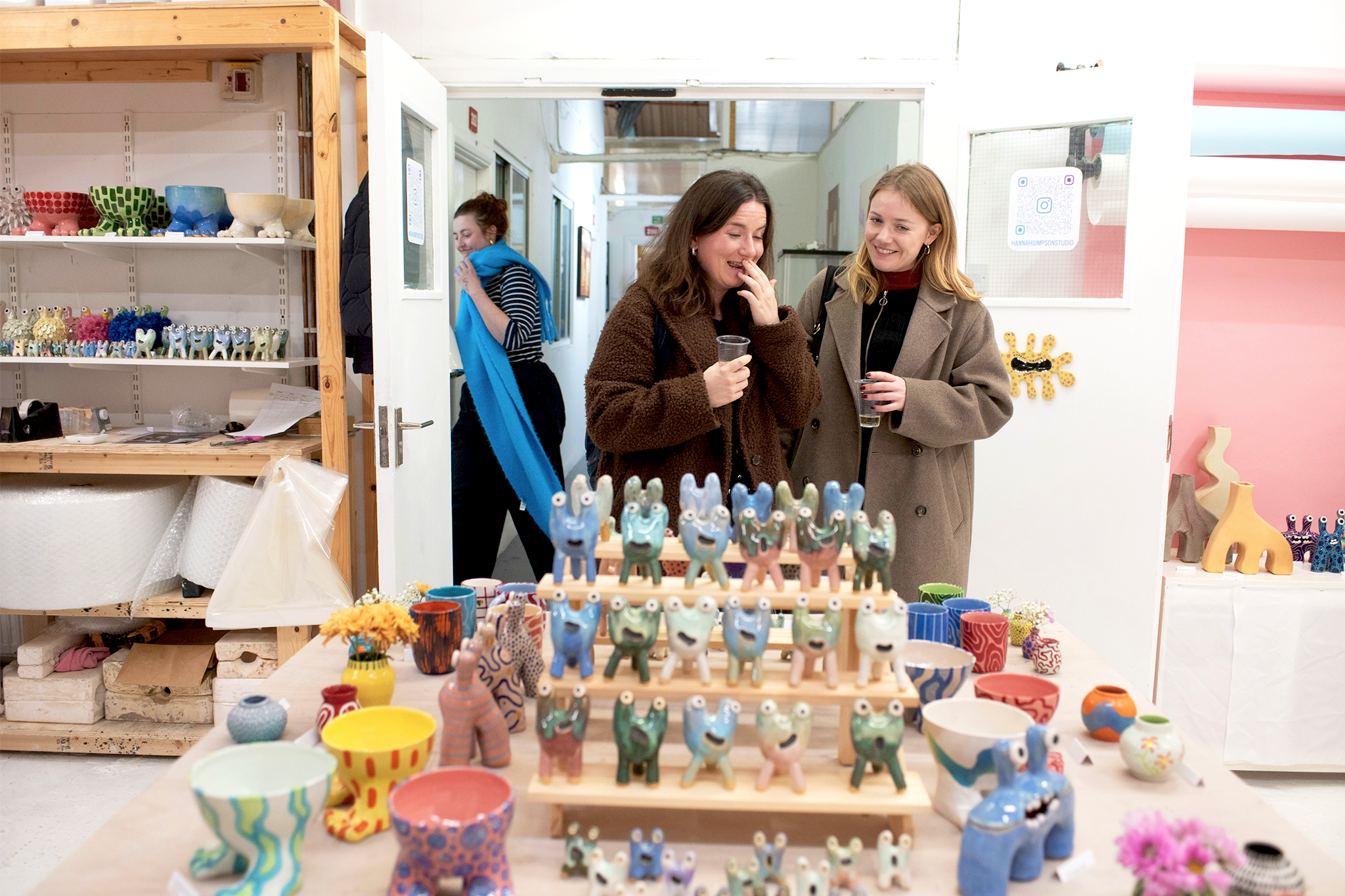 Two women browsing colorful ceramic animal figurines at an indoor craft market, with shelves of pottery and sculptures in the background.