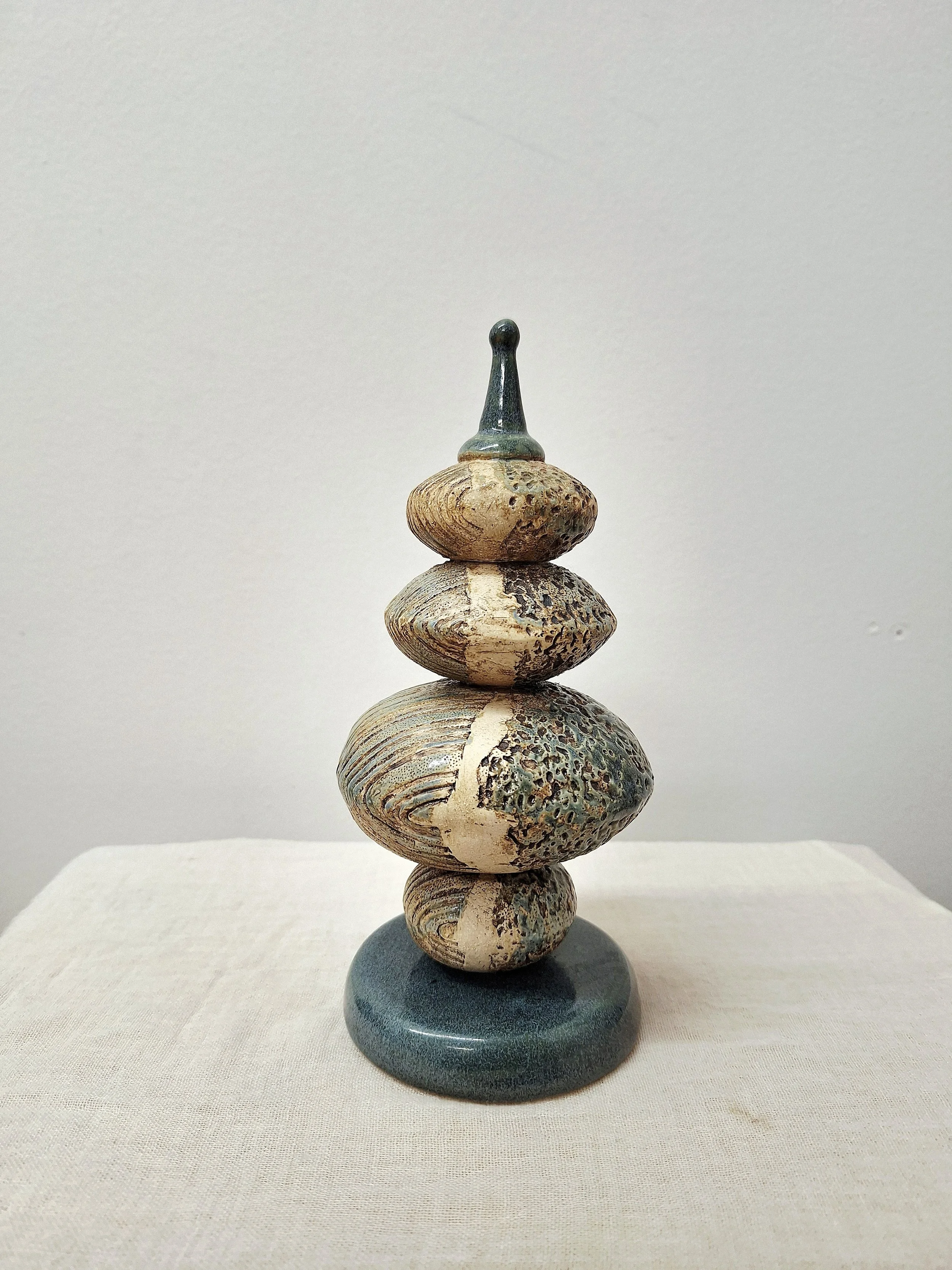 A sculpture made of stacked stones with a black, hand-shaped piece on top, displayed on a white pedestal on a wooden surface.