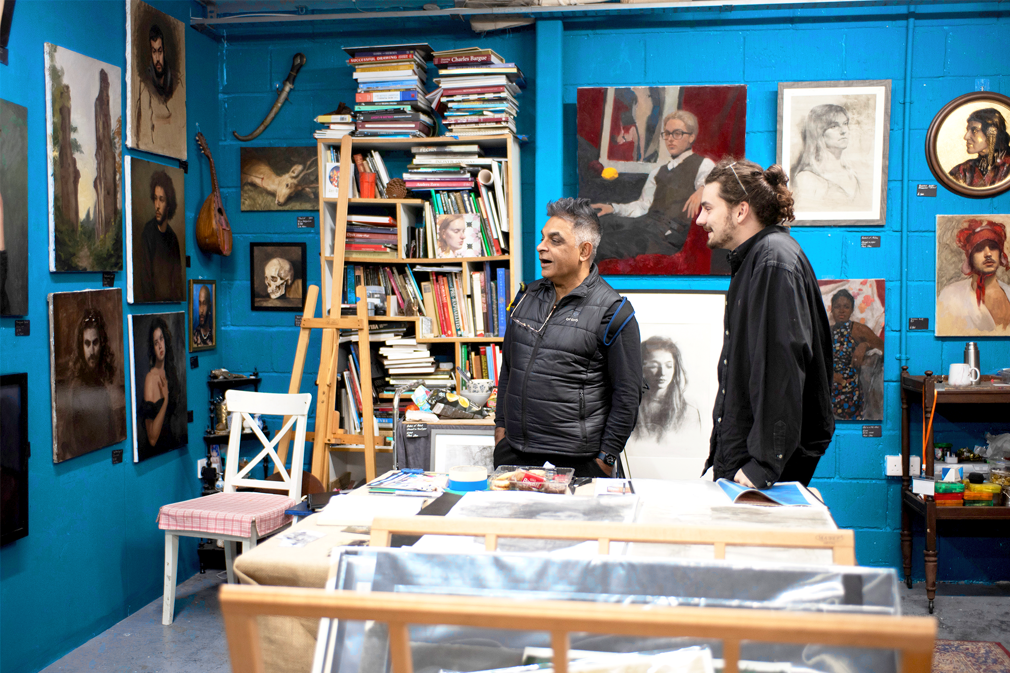 Two men are standing and talking in an art gallery with blue walls. The gallery is decorated with various portraits and artworks, some framed and others on canvases. There are shelves with books and art supplies in the background.