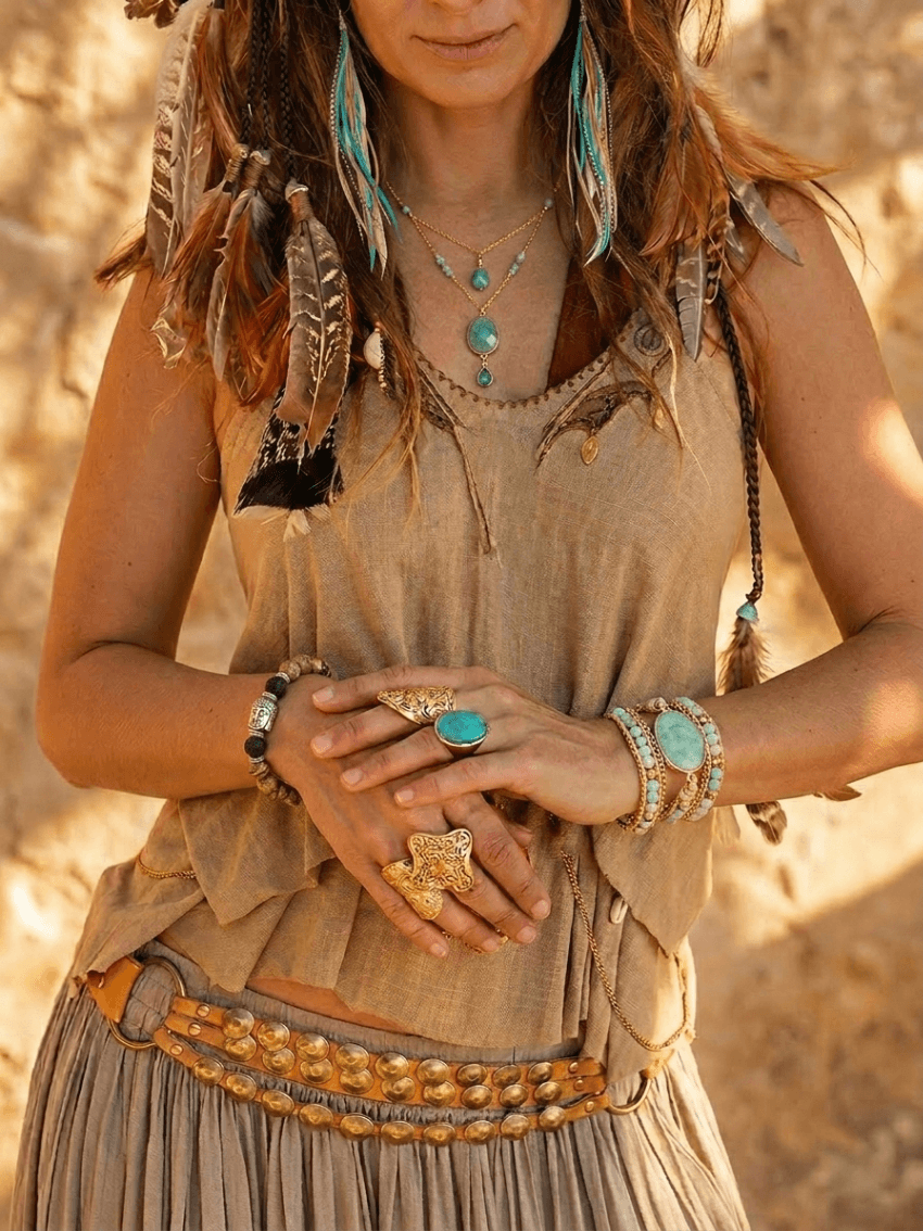 A woman wearing bohemian-style jewelry, including layered turquoise necklaces, rings, and bracelets, with feathers in her hair and dressed in earthy-toned clothing against a desert background.