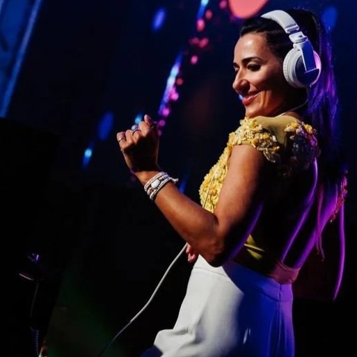 A woman DJ wearing headphones, smiling, and dancing at a nighttime event with colorful lights.