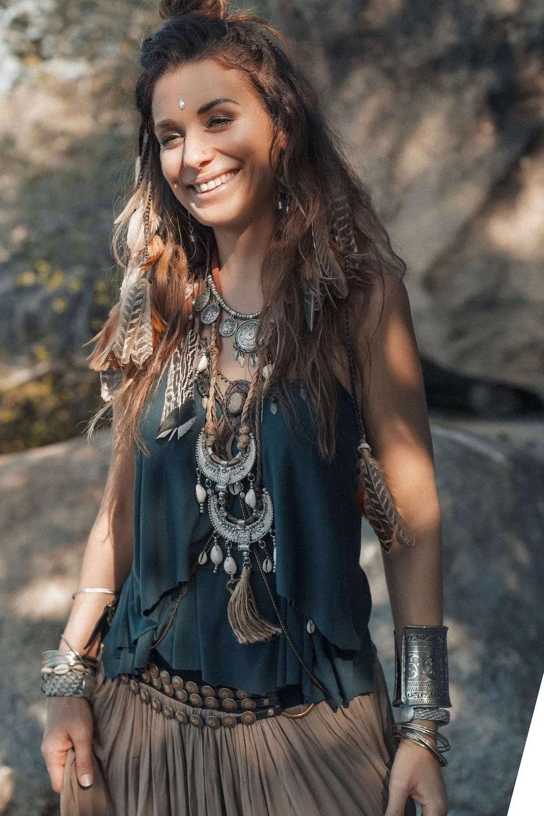 Woman with long, wavy hair wearing layered necklaces, bracelets, and bohemian clothing, smiling outdoors.