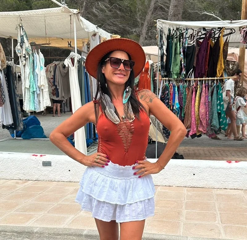 Woman wearing wide-brimmed hat, sunglasses, and summer outfit, standing in front of outdoor clothing market with racks of dresses and shirts.