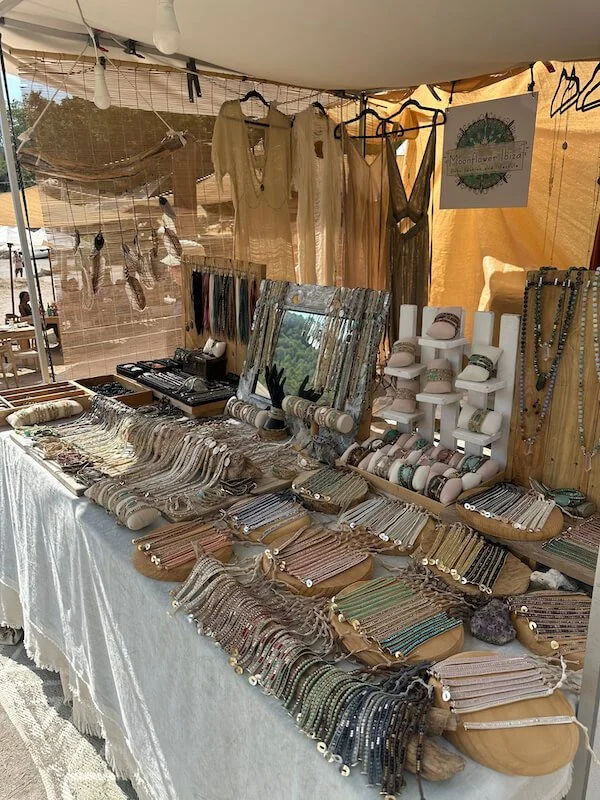 Booth at an outdoor market displaying jewelry, scarves, and clothing, with a mirror and a sign in the background.