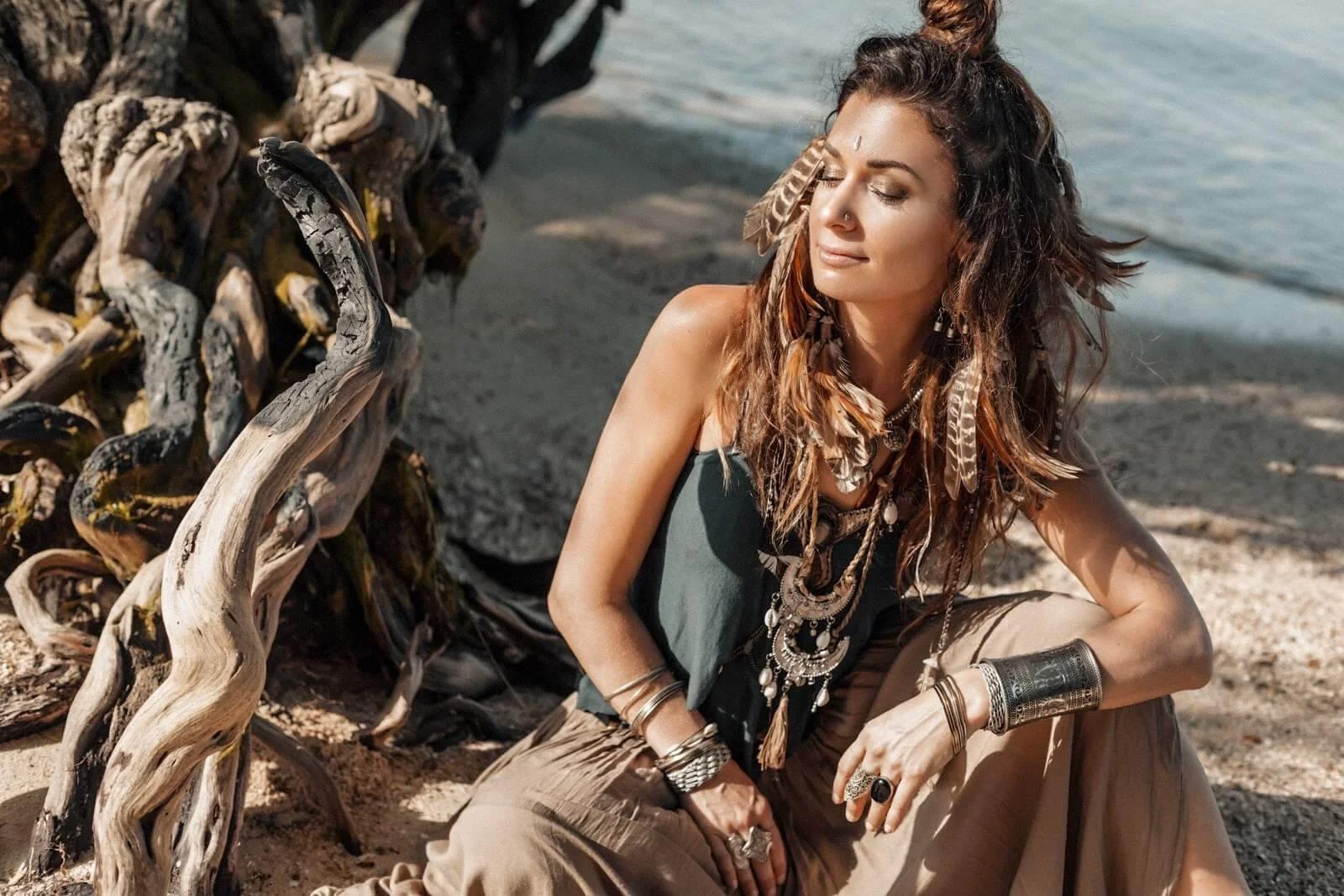 A woman with long, wavy hair adorned with feathers and jewelry, sitting on a beach near a piece of driftwood, with the ocean in the background.