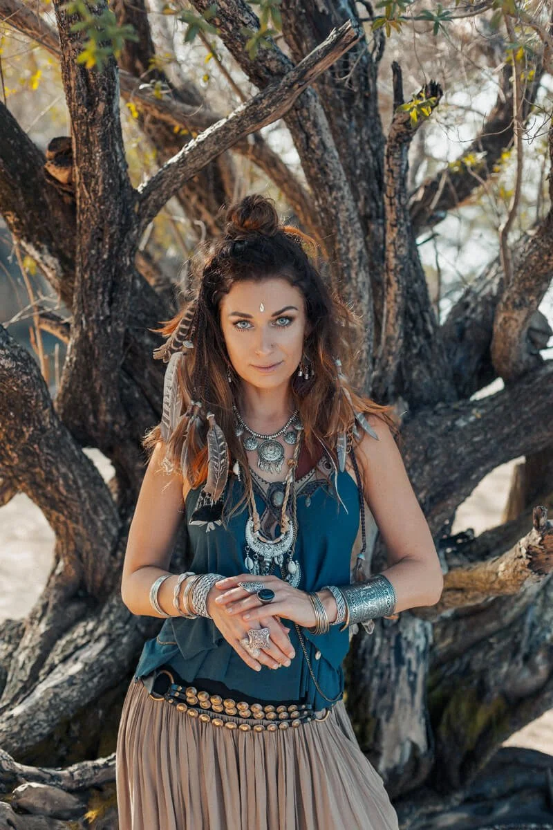 A woman with long, wavy brown hair wearing bohemian jewelry and clothing, standing in front of a large, gnarled tree outdoors.