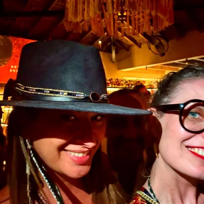 Two women posing for a photo in a dimly lit, rustic indoor setting. The woman on the left is wearing a large black hat decorated with a chain and has a broad smile. The woman on the right, partially visible, has glasses, red lipstick, and a cheerful expression.
