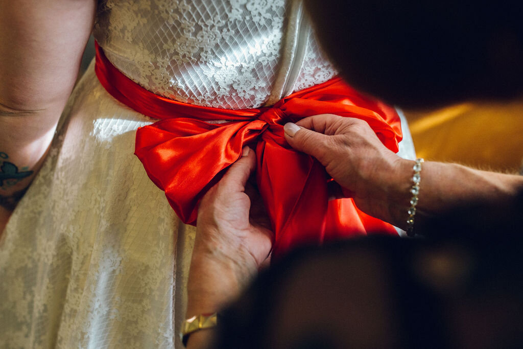 Red bow sash being tied on 50s wedding dress - Intimate Essex wedding Old Parish Rooms