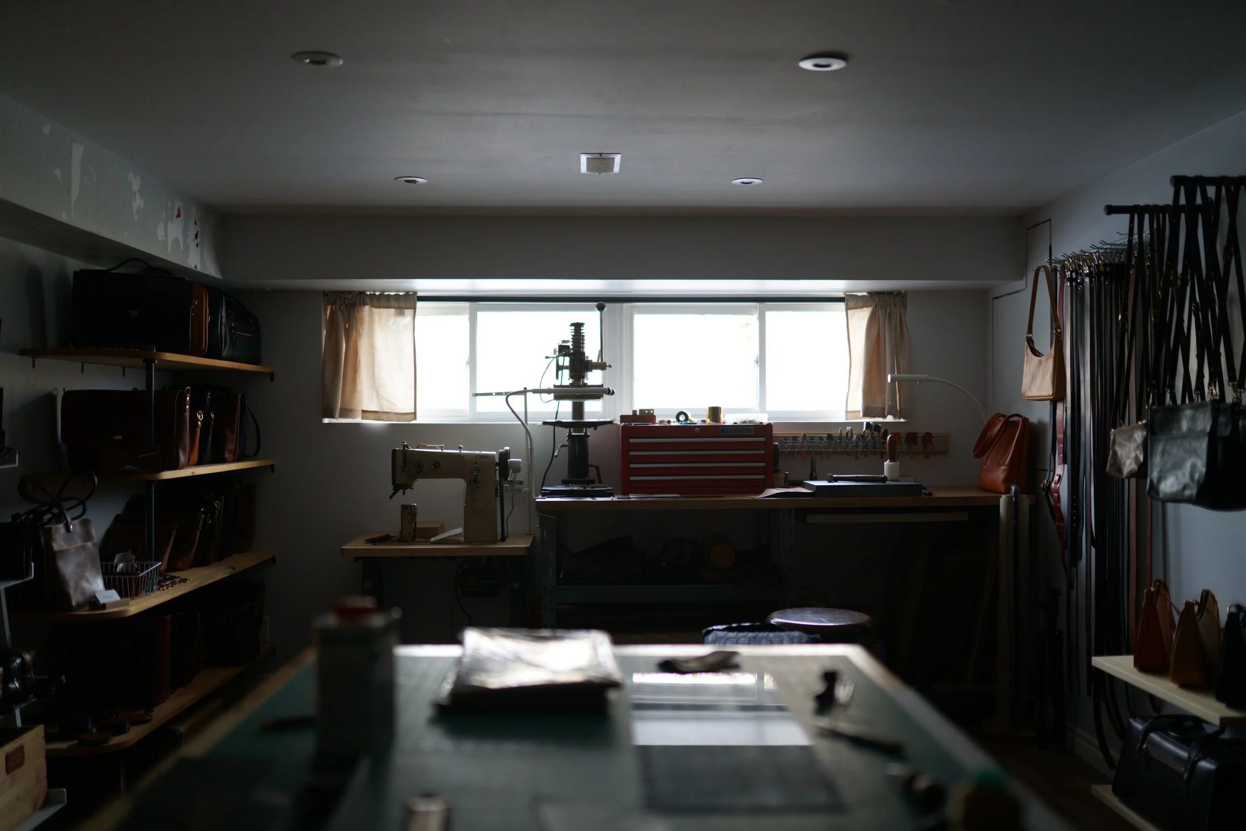 A dimly lit workshop with shelves of handbags on the left and right sides, a workbench with sewing tools in the middle, and a window with curtains at the back letting in natural light.