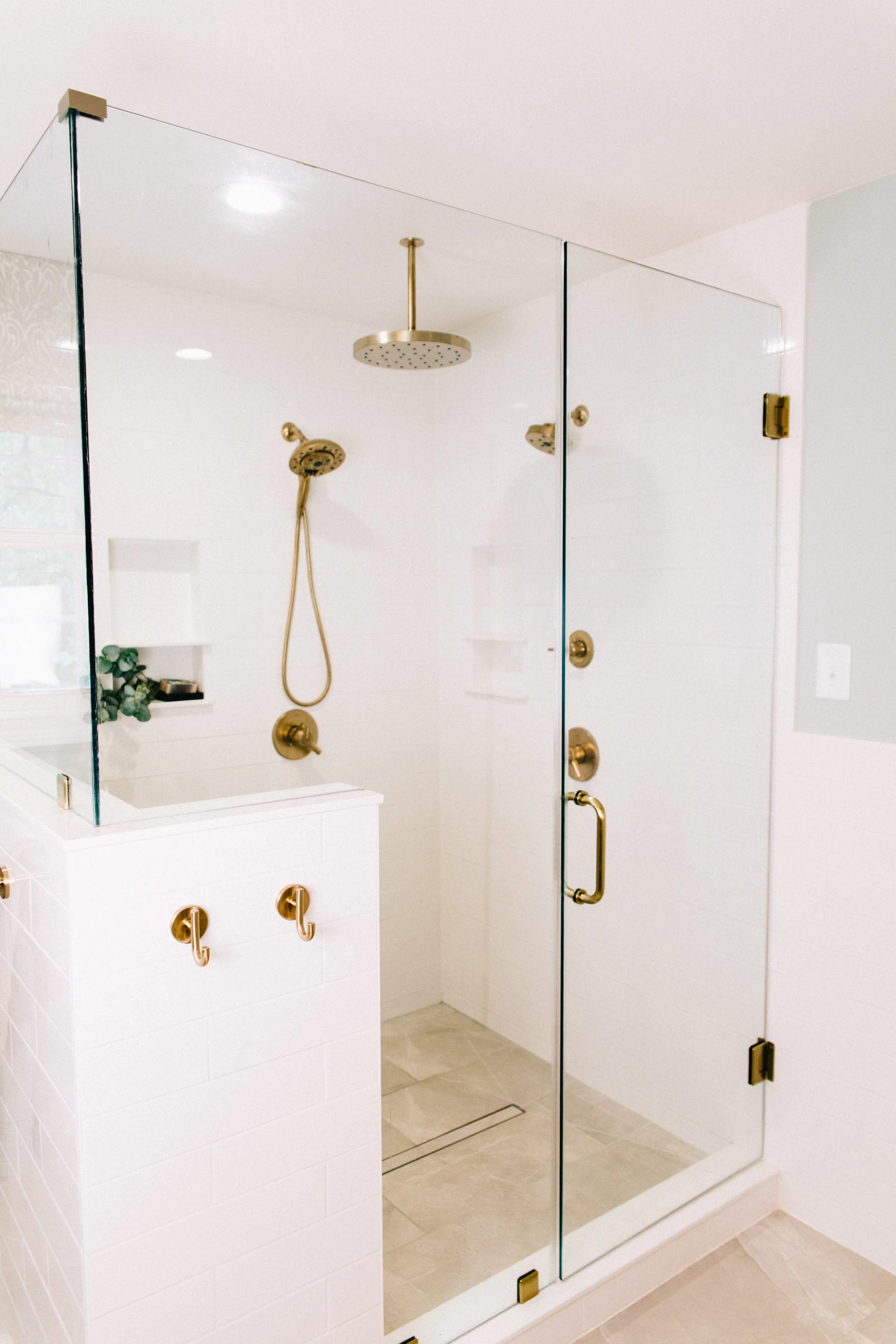 A modern, glass-enclosed shower with brass hardware, including a rainfall showerhead, a handheld shower, and several hooks on a white tiled wall, with neutral-toned flooring.
