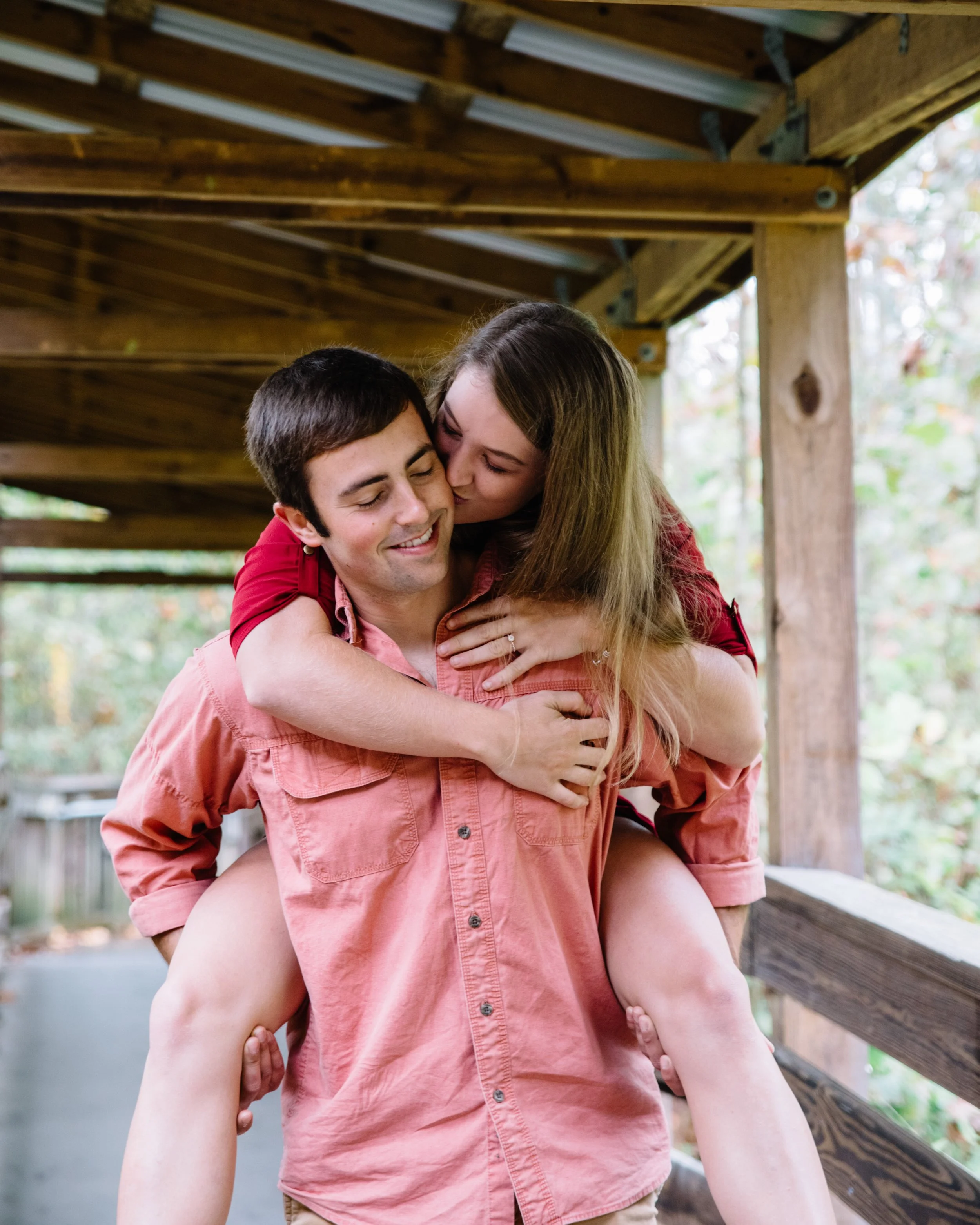 A couple sharing a happy piggyback ride outdoors, embracing and smiling at each other on a wooden walkway surrounded by trees.