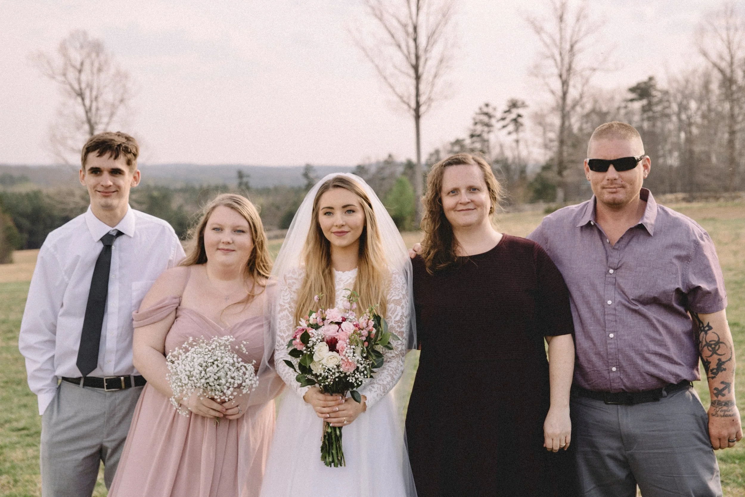 Group of five people outdoors, including a bride in a white lace wedding dress holding a bouquet, a bridesmaid in a pink dress holding a small bouquet, a man in a white shirt and black tie, a woman in a black dress, and a man in sunglasses and a purp