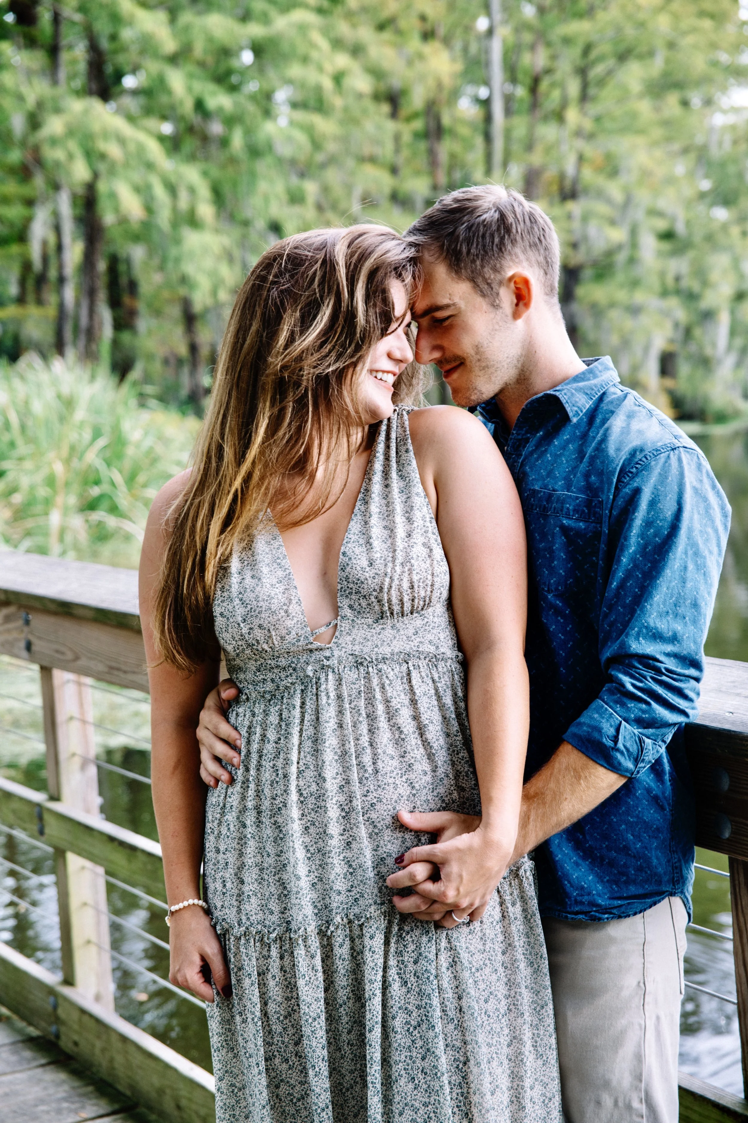 A young couple standing close together on a wooden bridge, smiling and touching foreheads in a natural, green outdoor setting.
