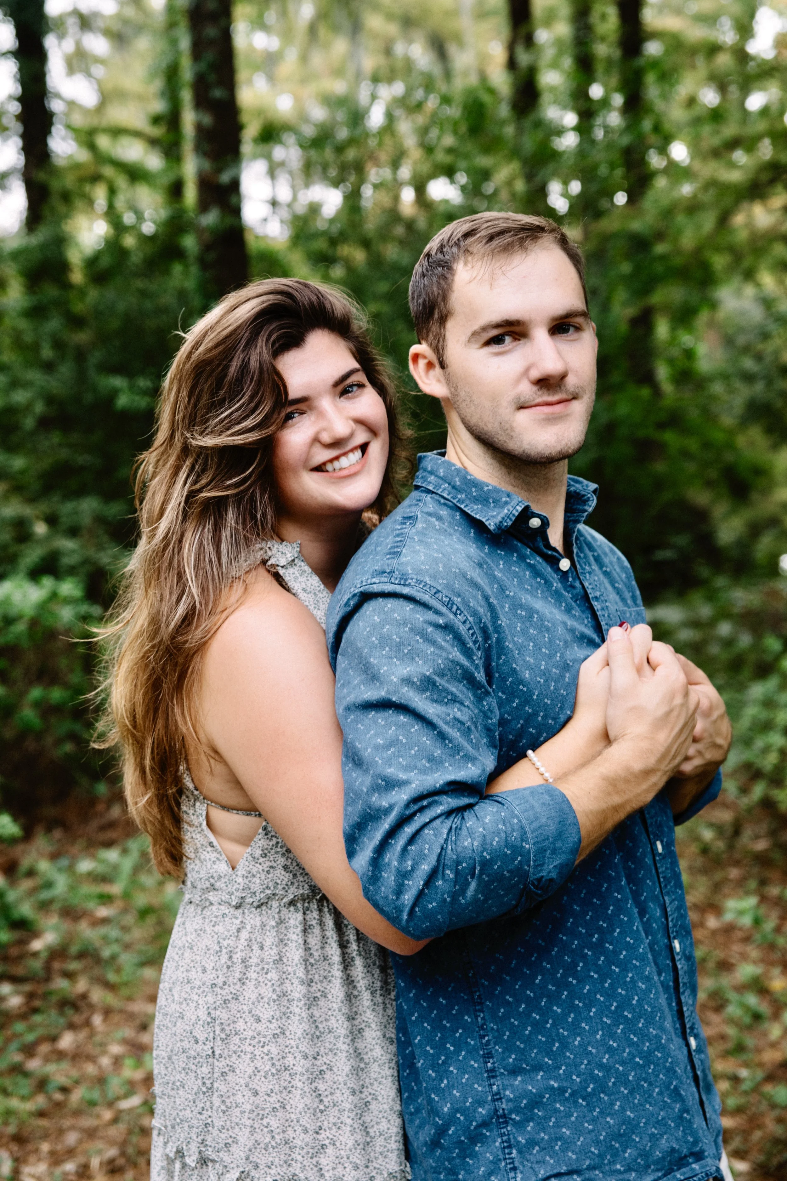 A young couple standing closely together outdoors in a wooded area, smiling at the camera, with the woman embracing the man from behind.