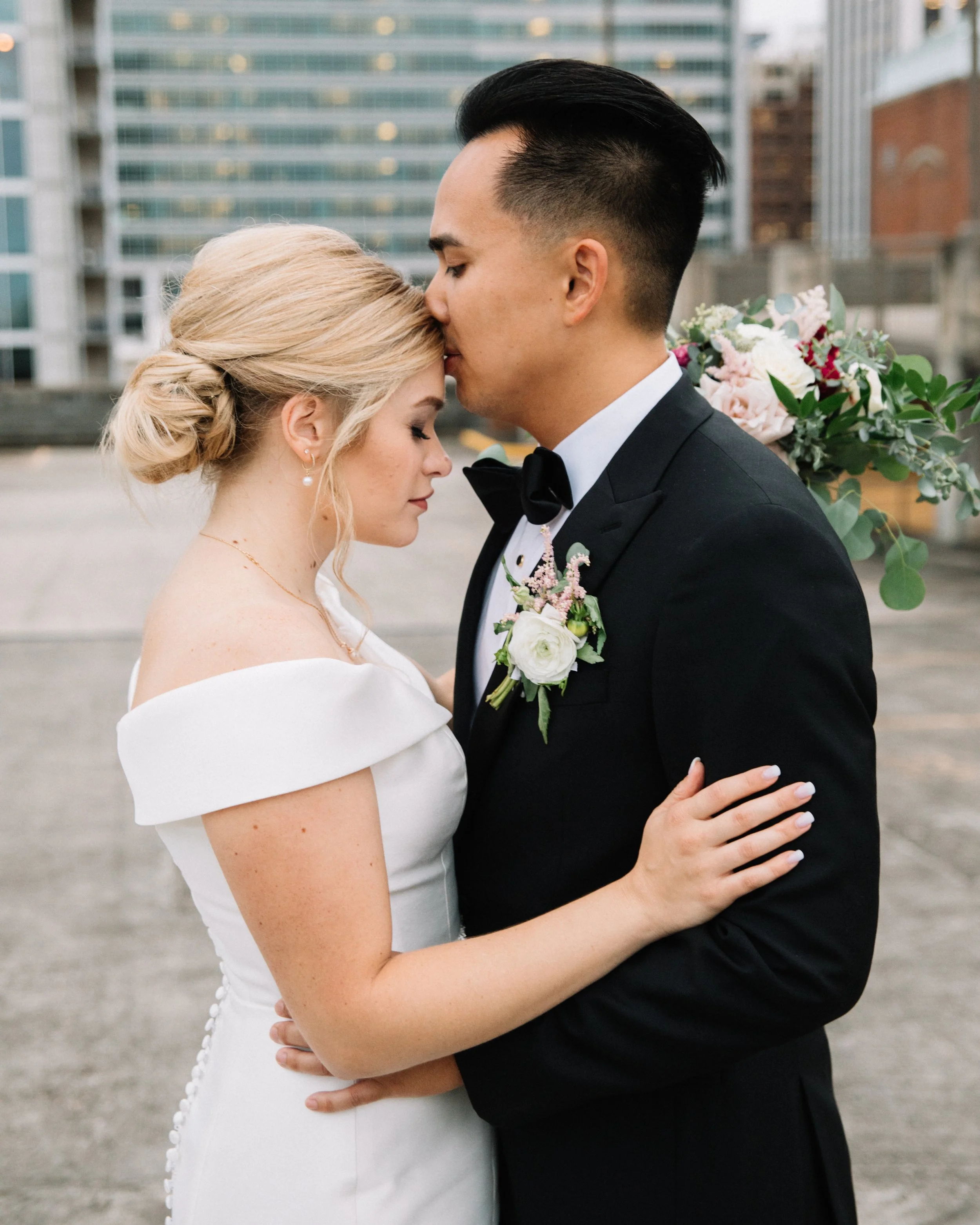 A newlywed couple embrace on a rooftop with city buildings in the background; the bride is in a white off-the-shoulder wedding dress with her hair in an elegant bun, and the groom is in a black tuxedo with a bow tie and boutonniere, tenderly touching