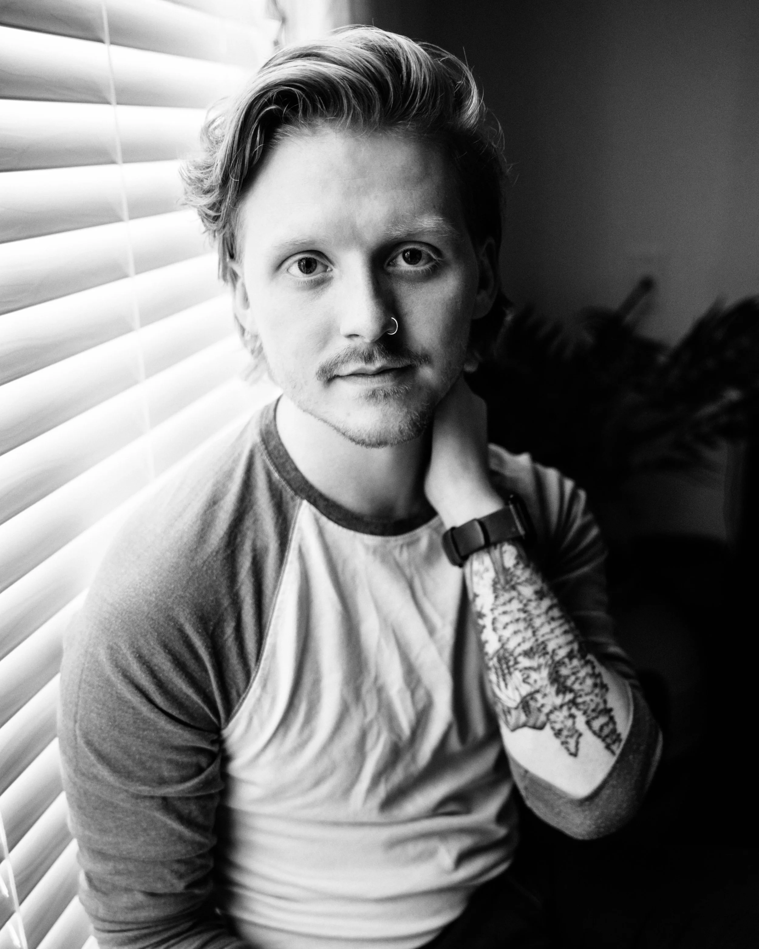 A young man with light skin, medium-length wavy hair, a nose ring, and a tattoo on his arm, sitting indoors near a window with blinds, looking into the camera.
