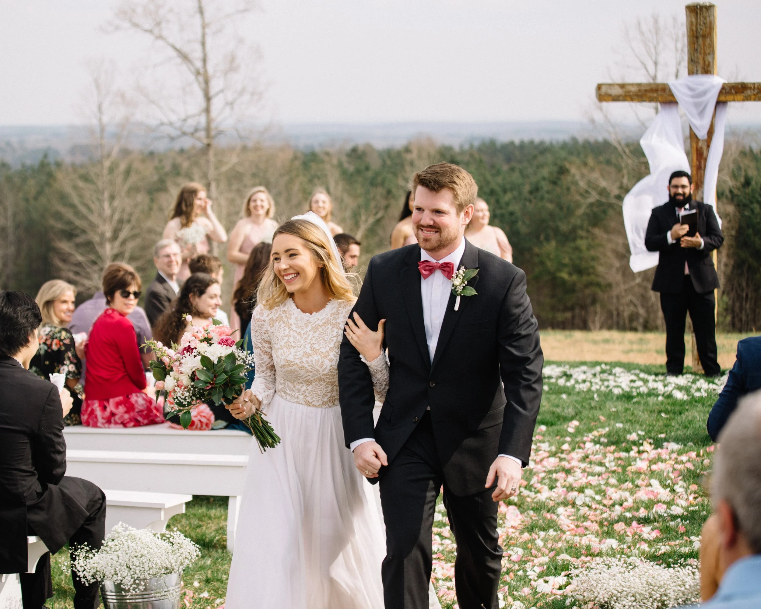 A bride and groom walking arm in arm down the aisle during an outdoor wedding ceremony, surrounded by seated guests and a rustic wooden cross draped with white fabric, in a natural setting with trees in the background.