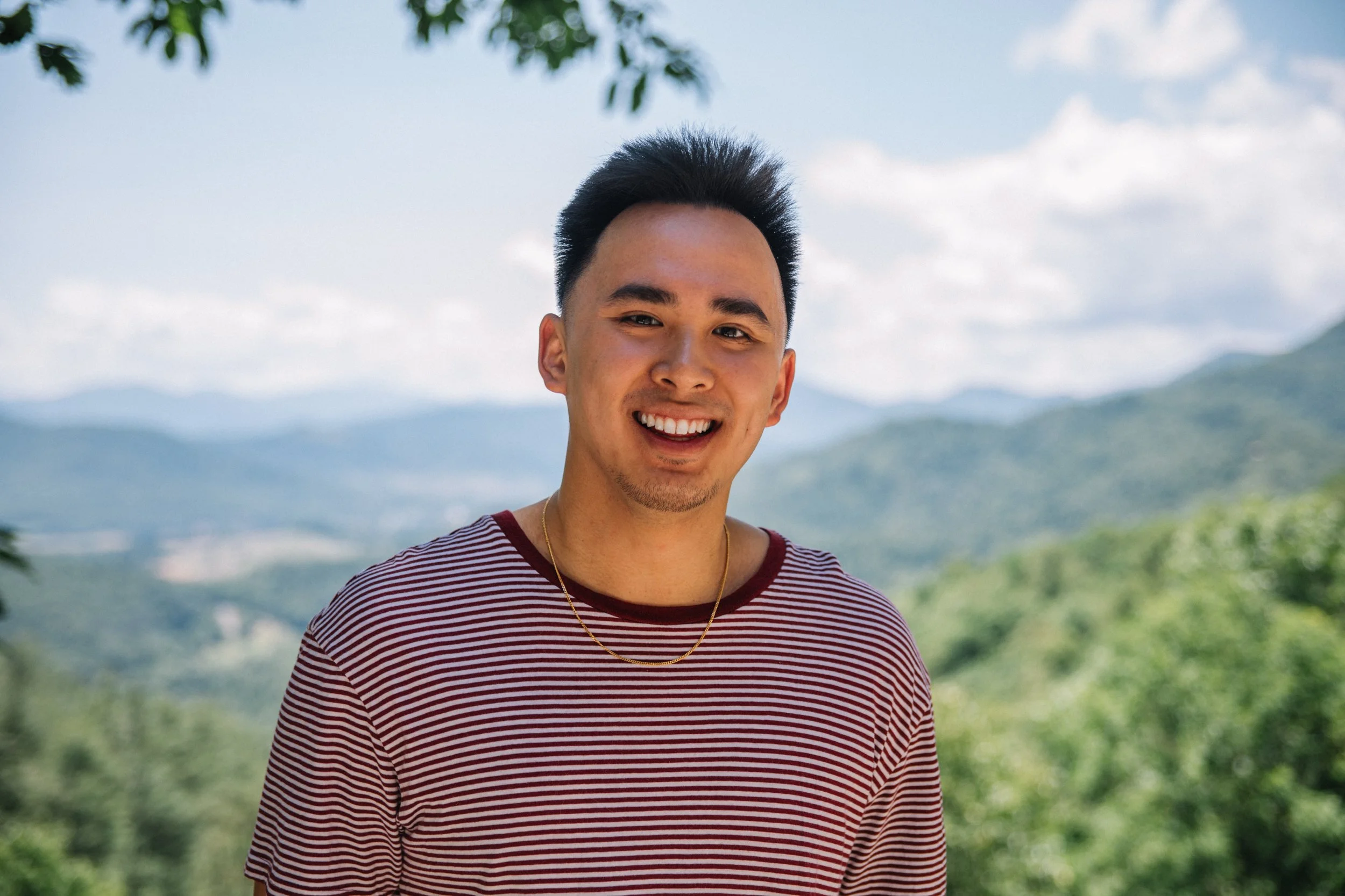 A smiling young man with black hair, wearing a red and white striped shirt and a gold chain, standing outdoors with a background of mountains and blue sky.