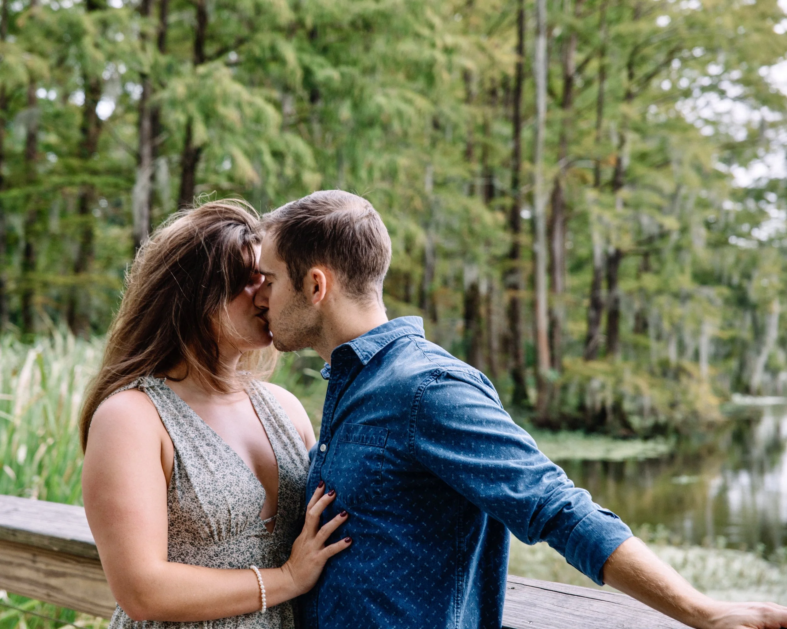 A couple kissing outdoors near a body of water and trees.