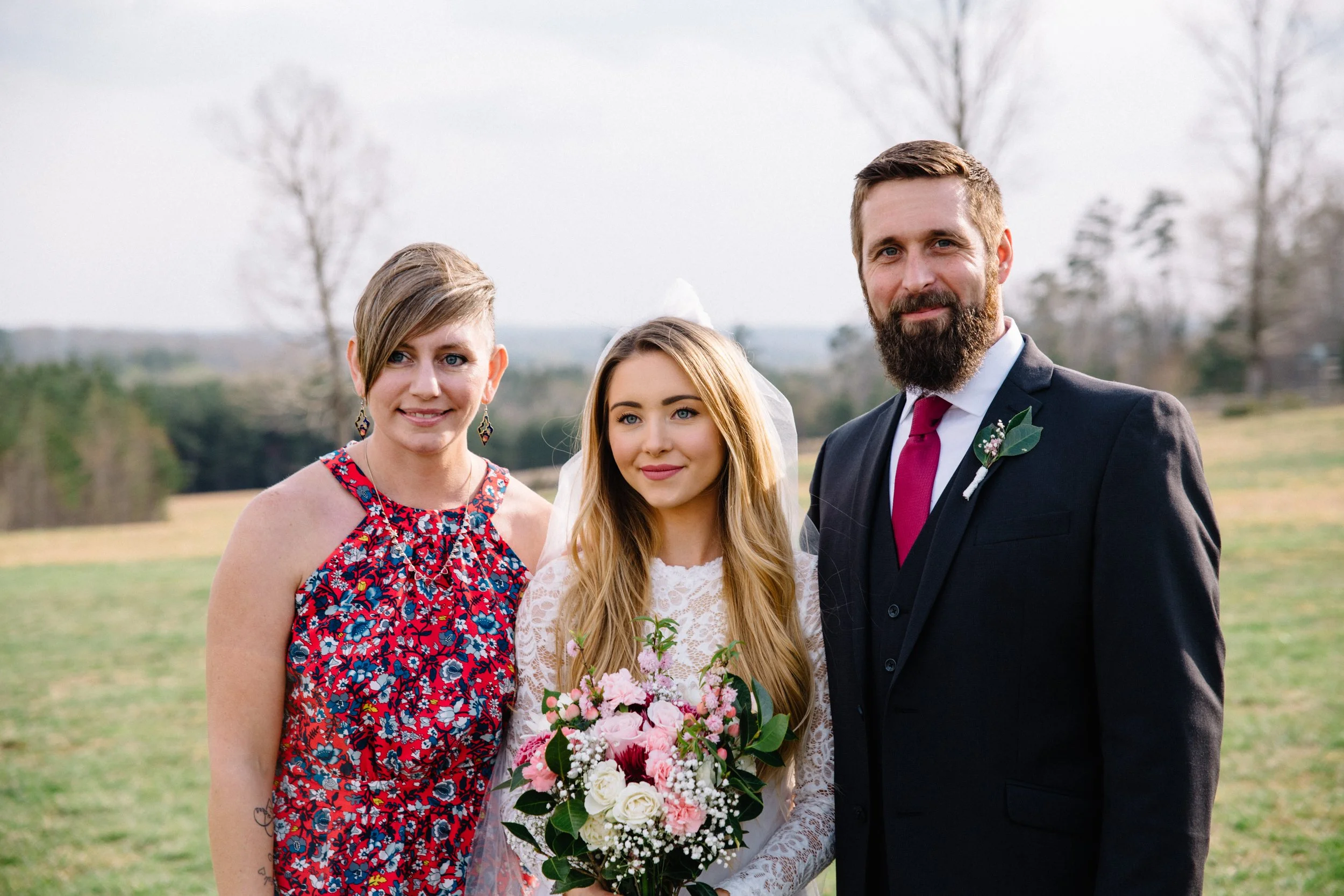 A bride holding a bouquet of pink and white flowers, standing between a woman in a colorful floral dress and a man in a black suit with a red tie, in an outdoor field with trees in the background.