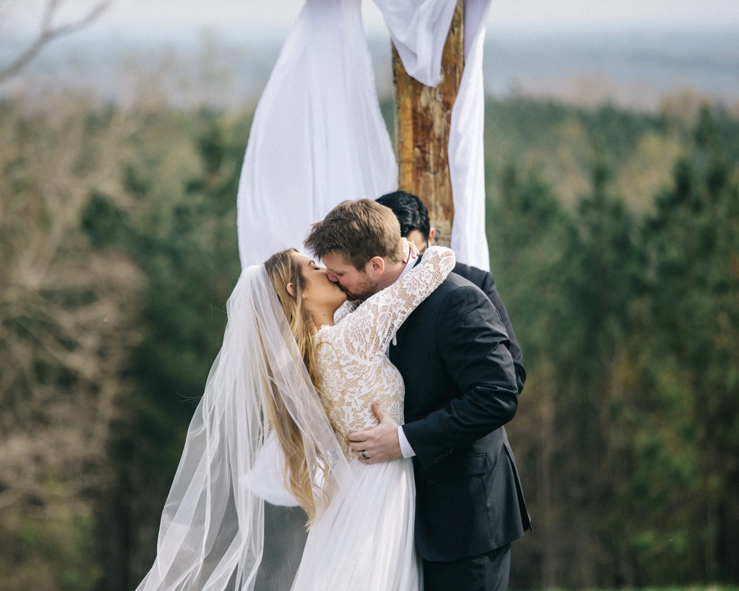 A bride and groom share a kiss during their outdoor wedding ceremony, standing beneath a wooden arch decorated with white fabric, with a natural background of trees and greenery.