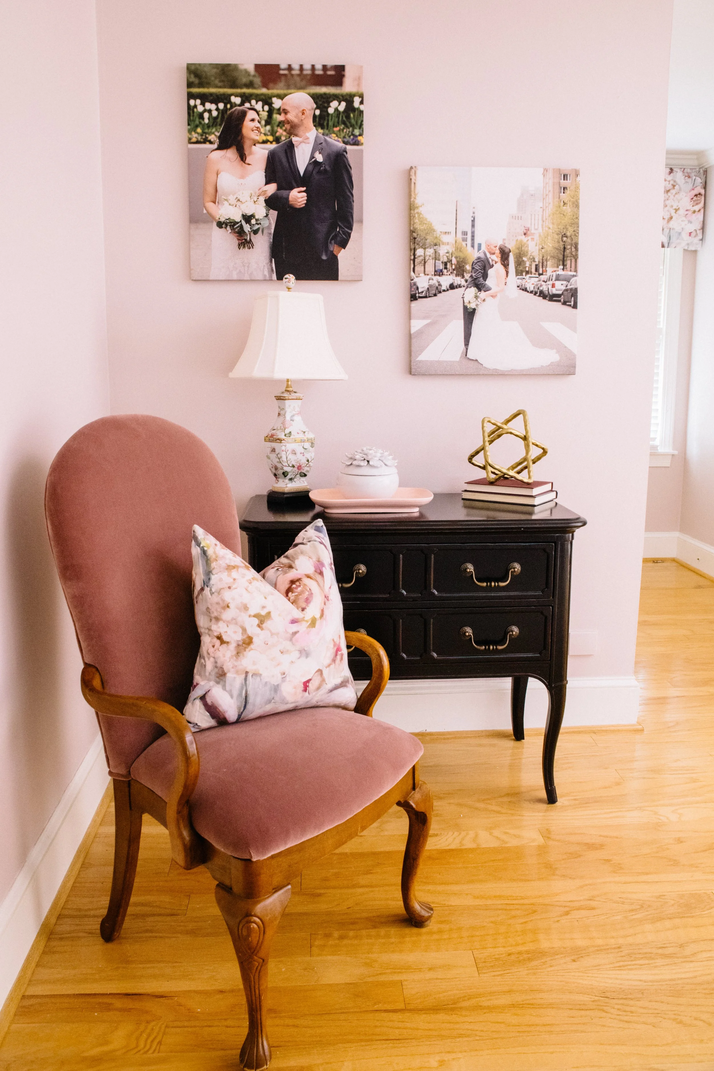 A pink room with a pink velvet armchair with a floral pillow and a black dresser with decorative items, framed wedding photos on the wall, a table lamp, and a window with floral curtains.