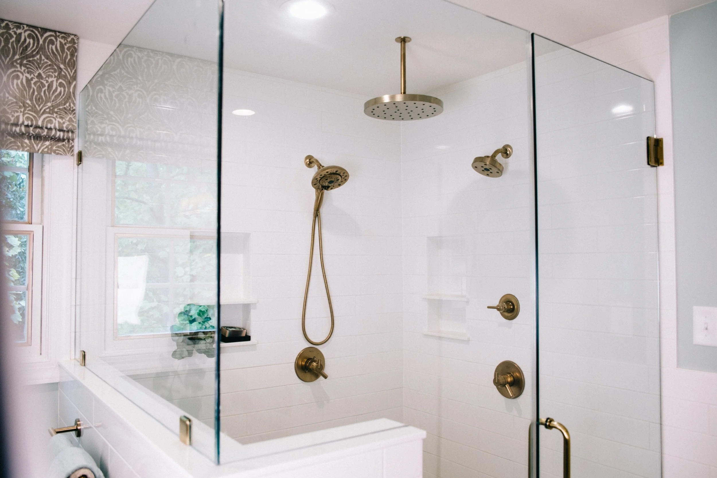 A bathroom shower with a glass enclosure, two brass showerheads, a handheld shower, and white tiled walls. There is a window with a patterned roller blind.