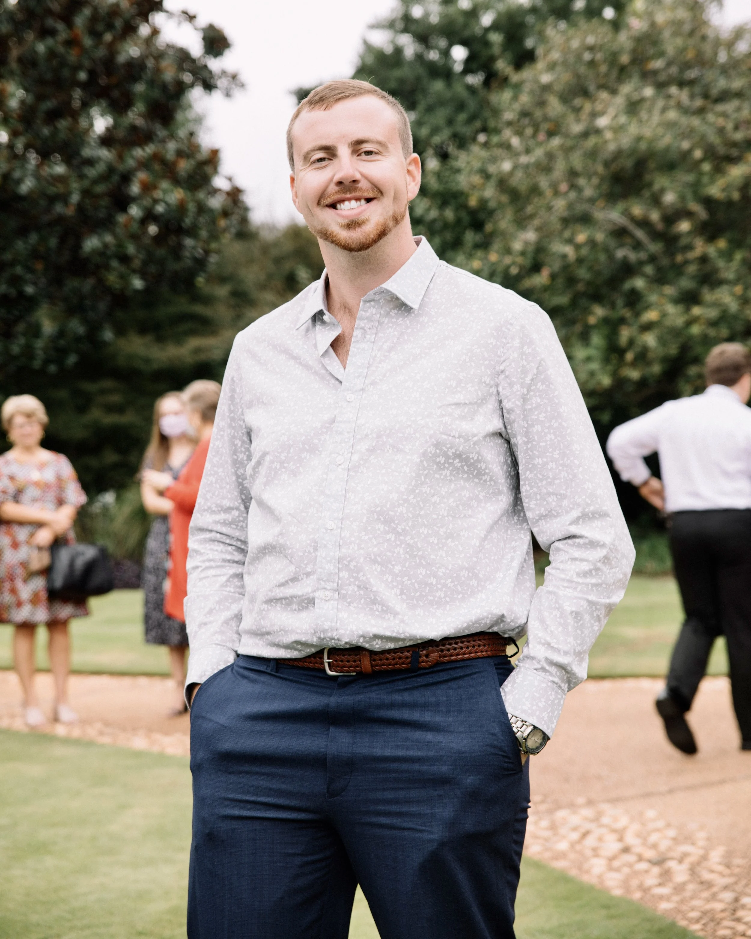 A smiling man in a white patterned shirt and navy blue pants standing outdoors with people in the background.