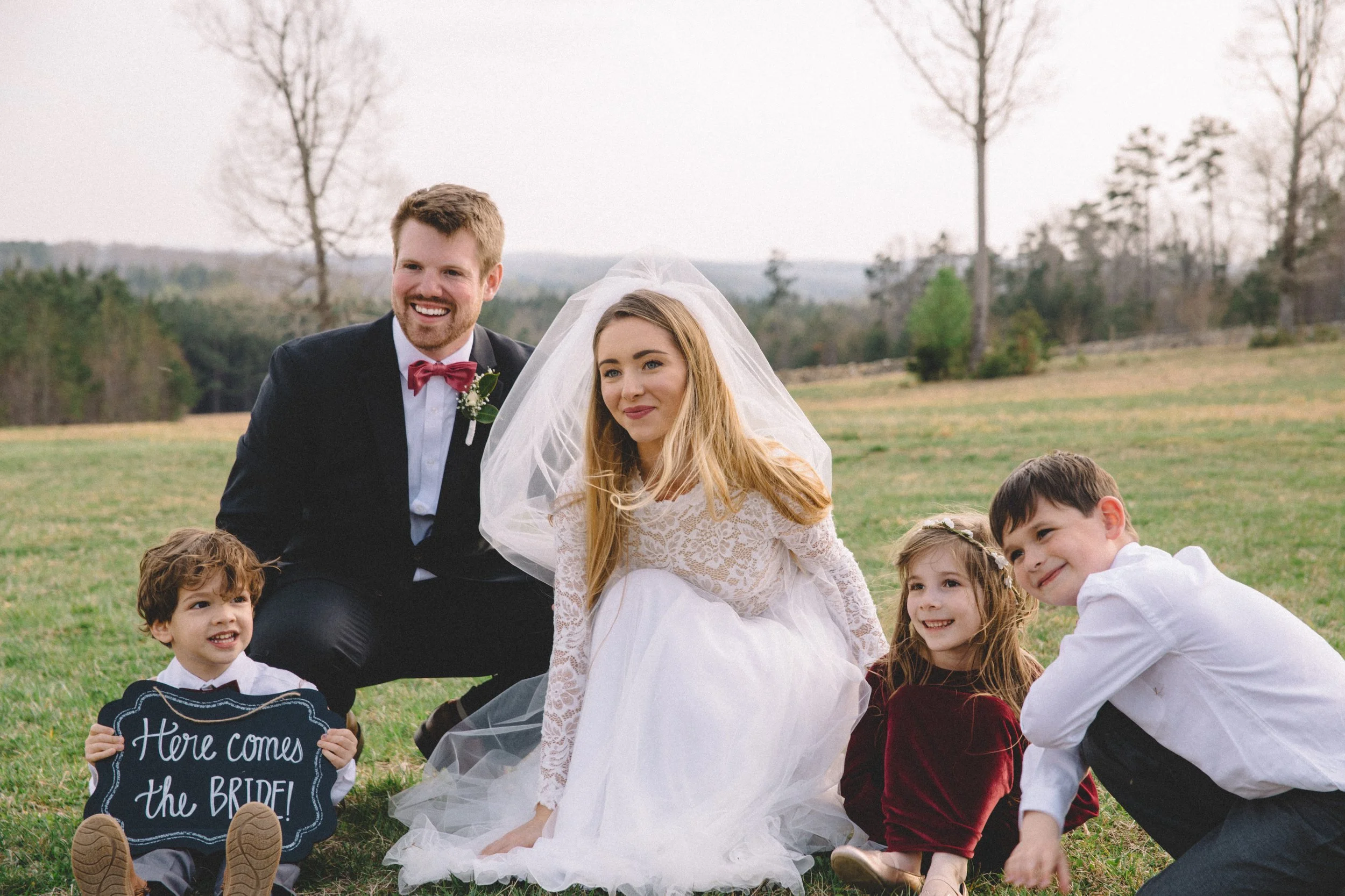 A wedding scene outdoors with a bride in a white lace dress and veil, a groom in a black suit with red bow tie, and three children sitting and lying on grass, one holding a sign that says "Here comes the BRIDE!" The background has a field with trees 