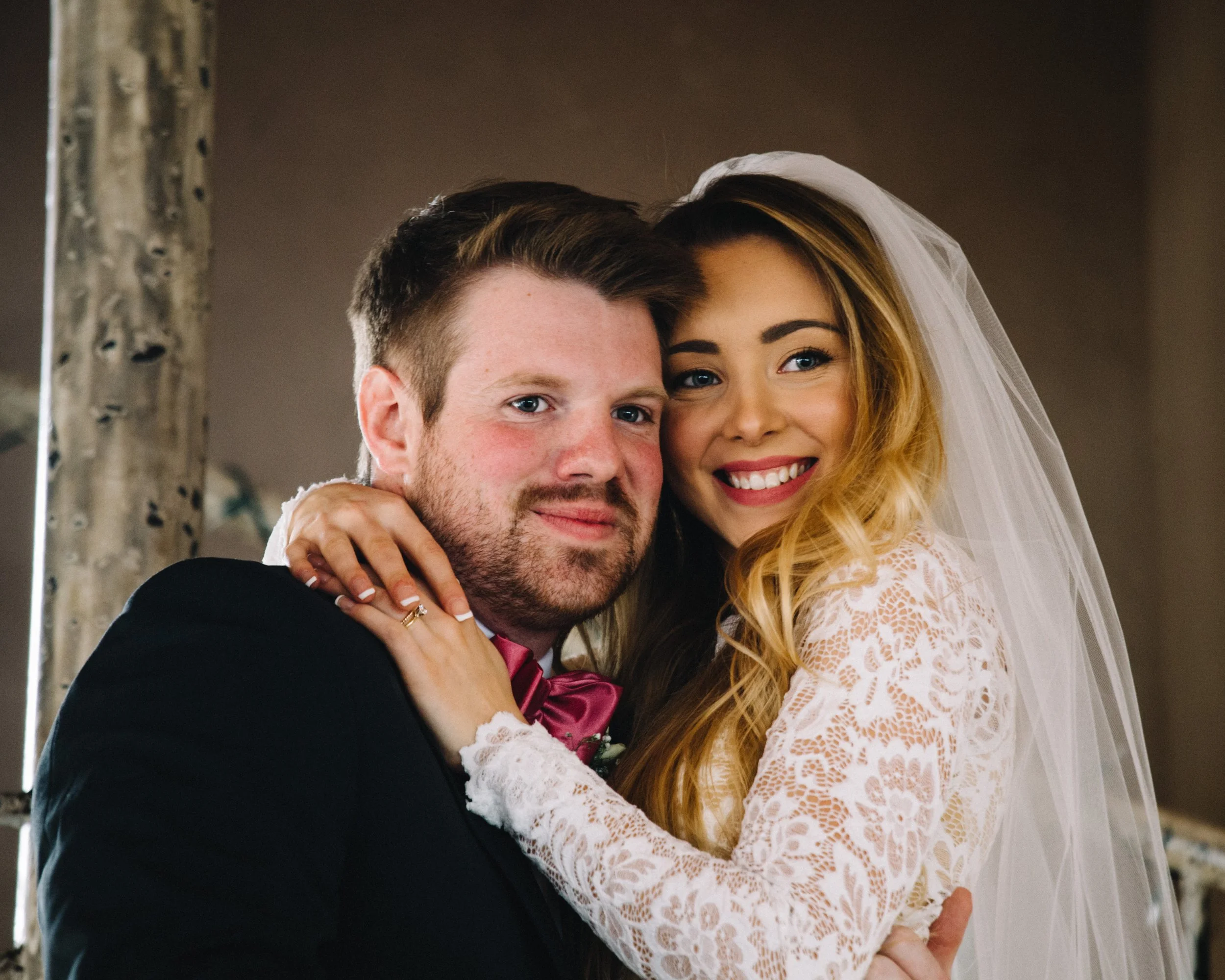 A bride and groom smiling and embracing each other on their wedding day. The bride has long, wavy blonde hair, a veil, and a lace wedding dress. The groom has short hair, a beard, and is wearing a black tuxedo with a bow tie.