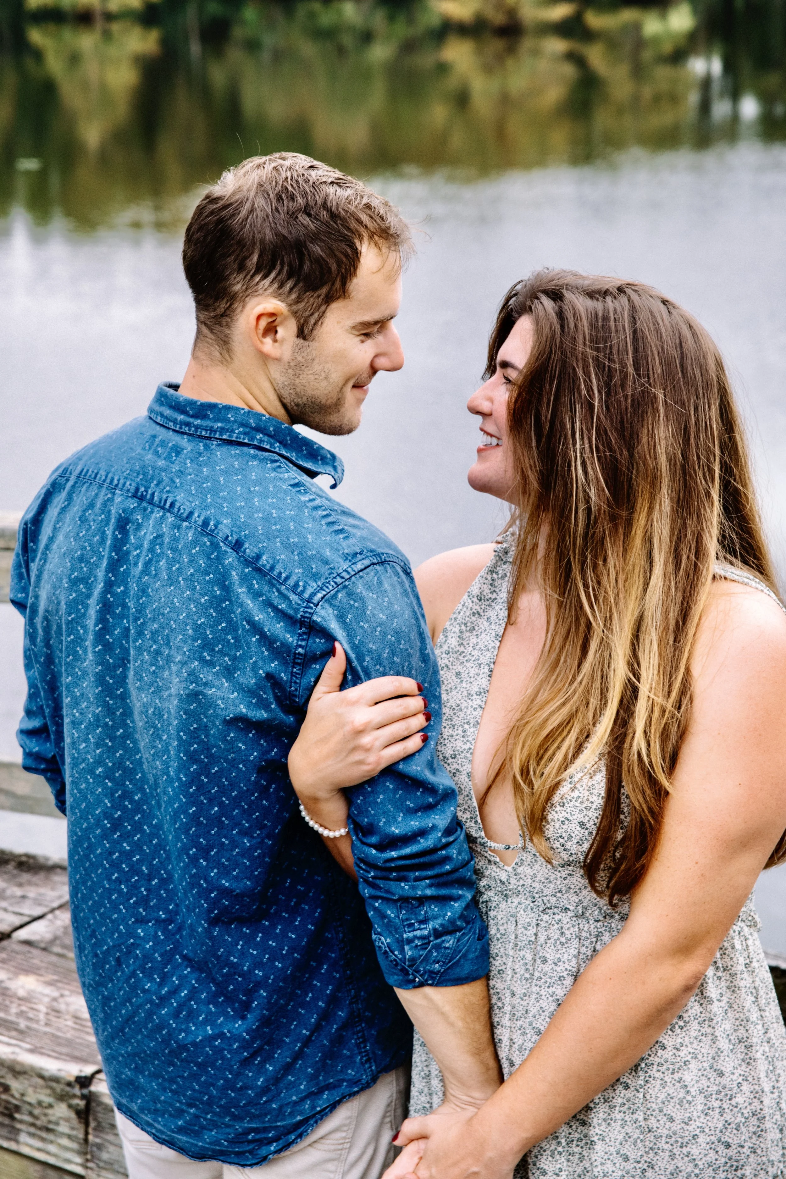 A couple standing close together outdoors by a lake, looking into each other's eyes and smiling.