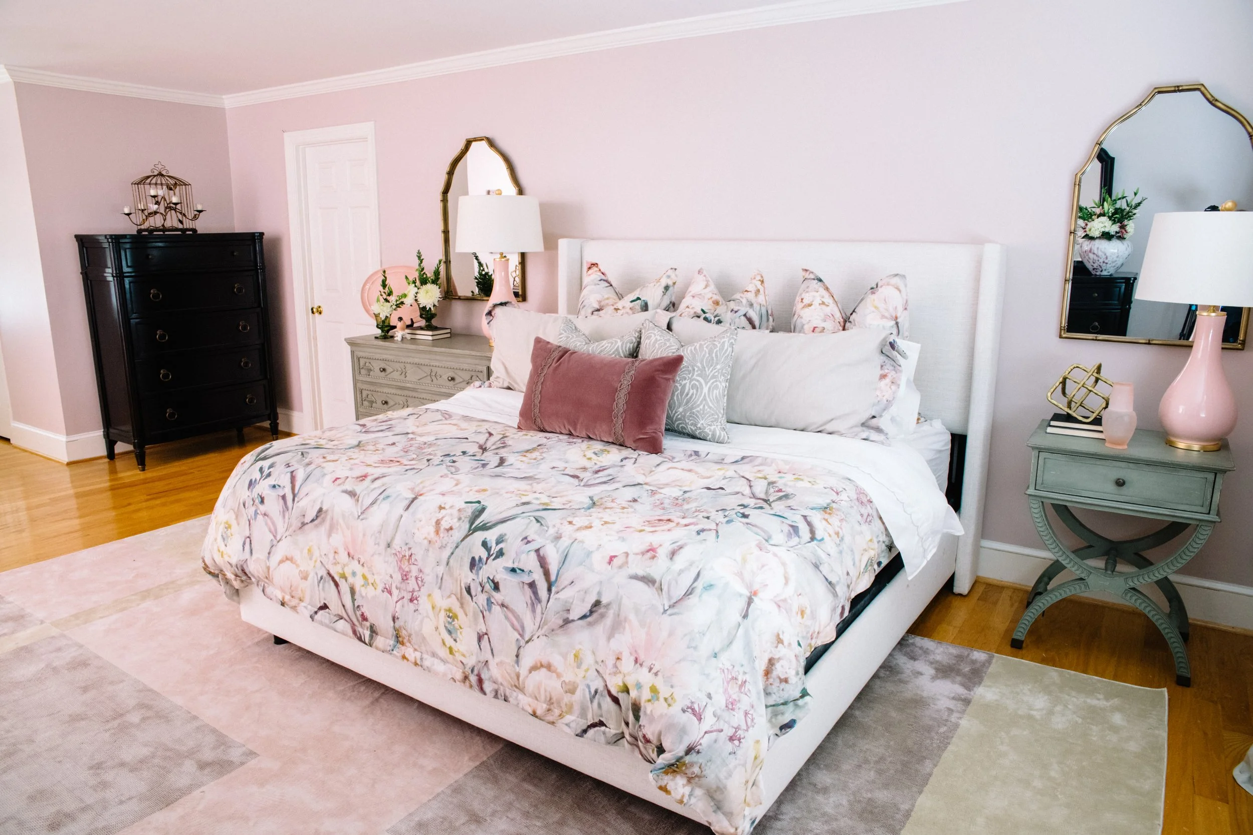 A bedroom with a large bed covered in floral bedding and multiple pillows, two nightstands with lamps, one black chest of drawers, and two mirrors on the pastel-colored walls.