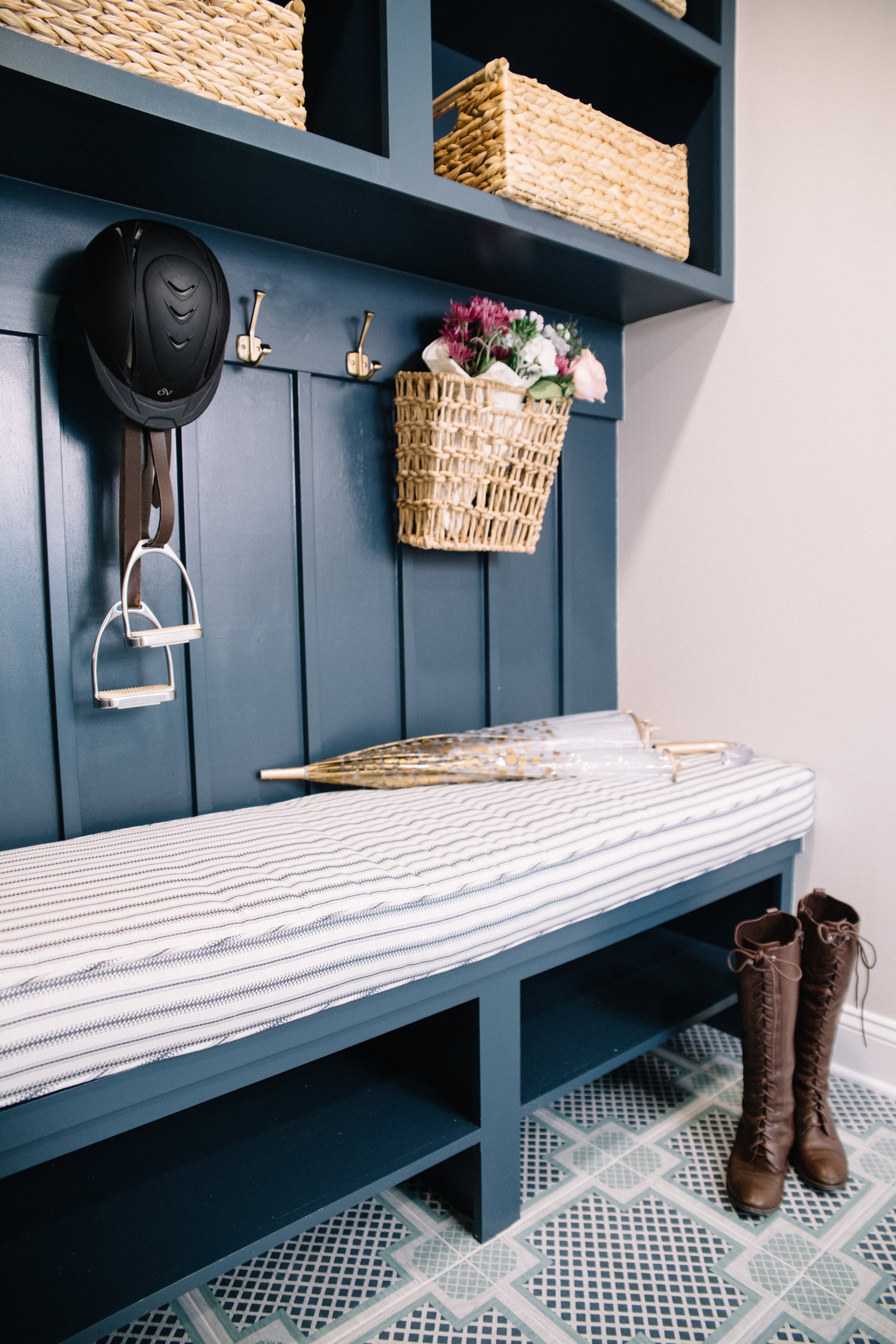 Blue entryway bench with a striped cushion, a pair of brown knee-high lace-up boots, a woven umbrella, a wall-mounted black helmet, a small basket of flowers, and a mint green tiled floor with a diamond pattern.