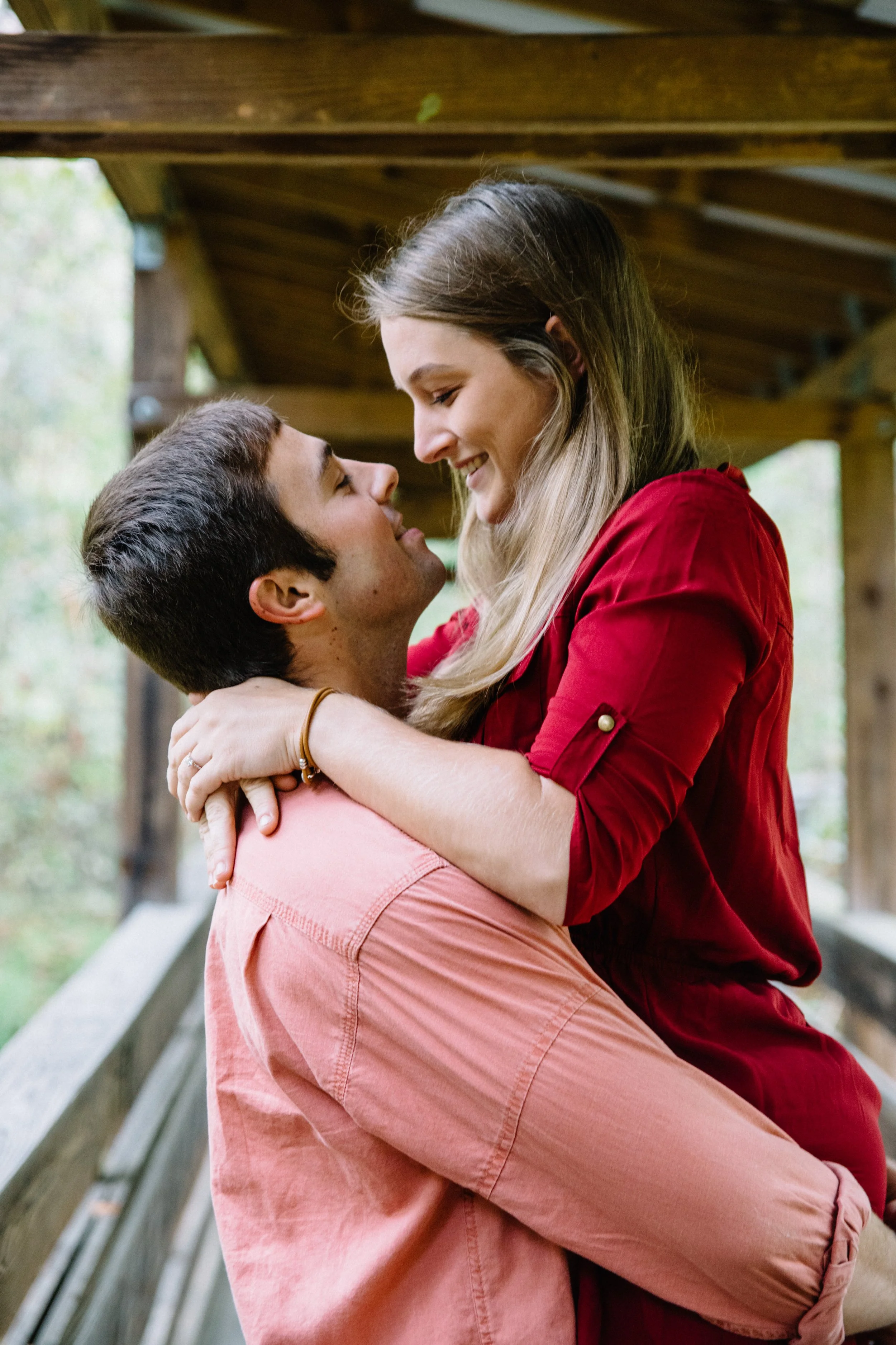 A couple is embracing on a wooden bridge, smiling at each other in a scenic outdoor setting.