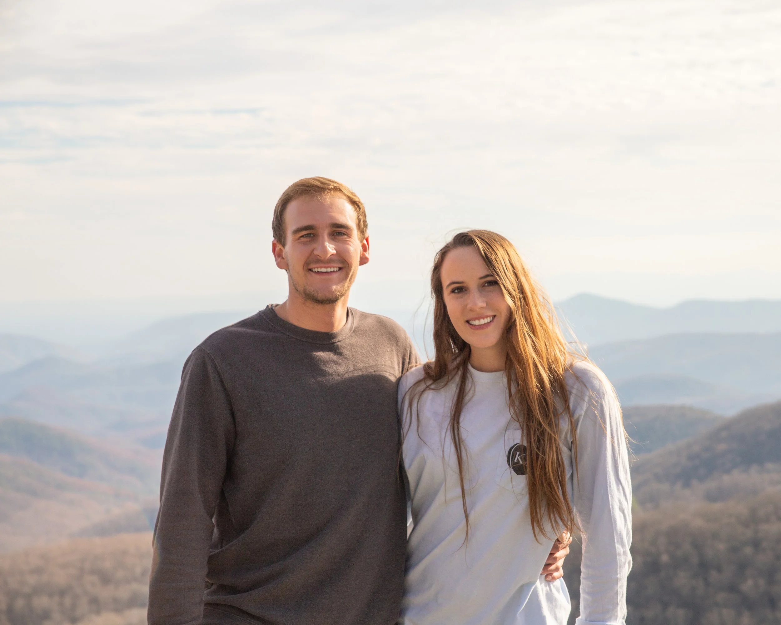 A smiling young man and woman standing outdoors on a mountain overlook, with rolling hills and a cloudy sky in the background.
