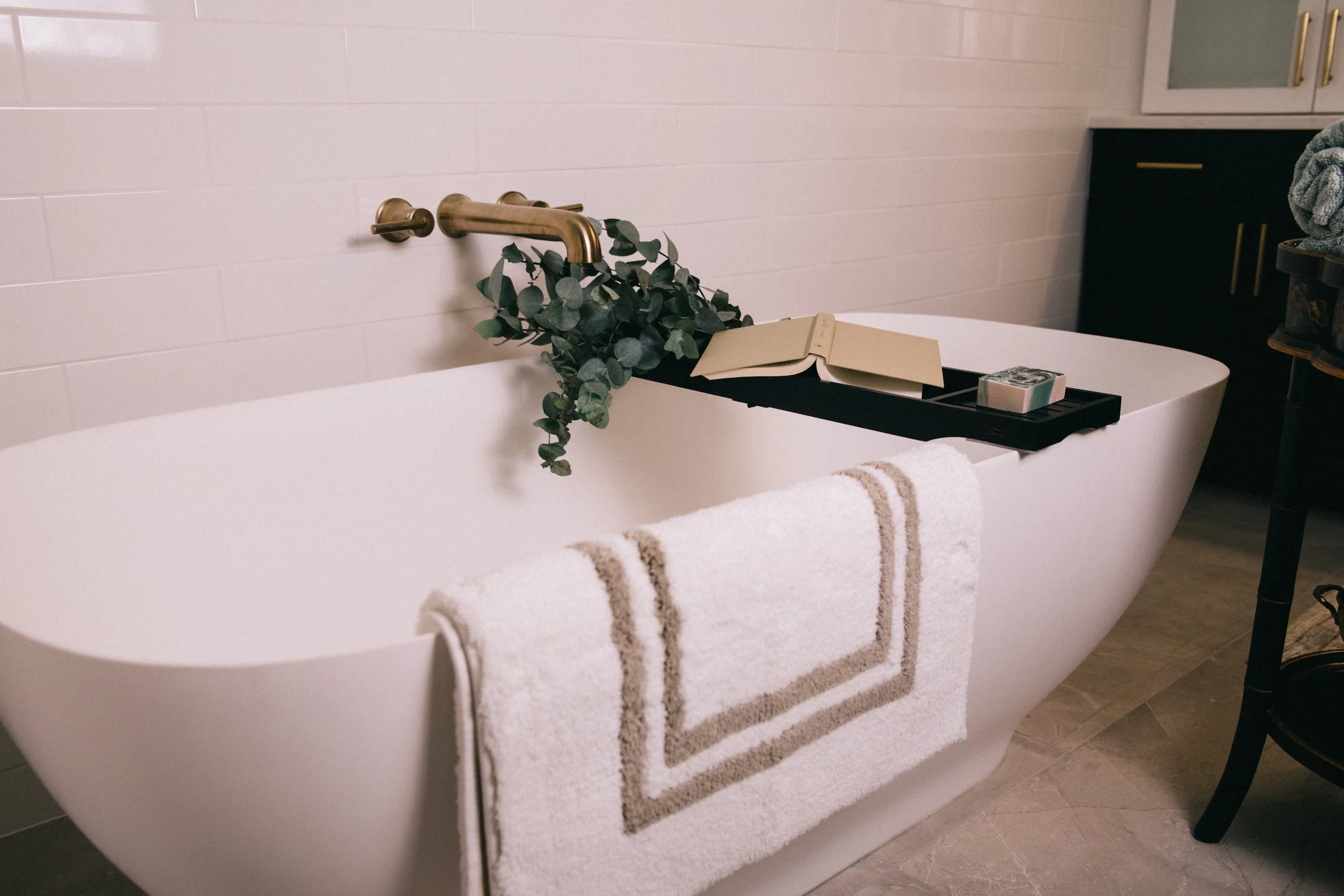 A white freestanding bathtub with a beige and brown striped towel hanging over the edge, a black tray holding an open book and a box of tissues, a green leafy plant on the tray, and a hand towel hanging on the side. There is a brass faucet on the wal