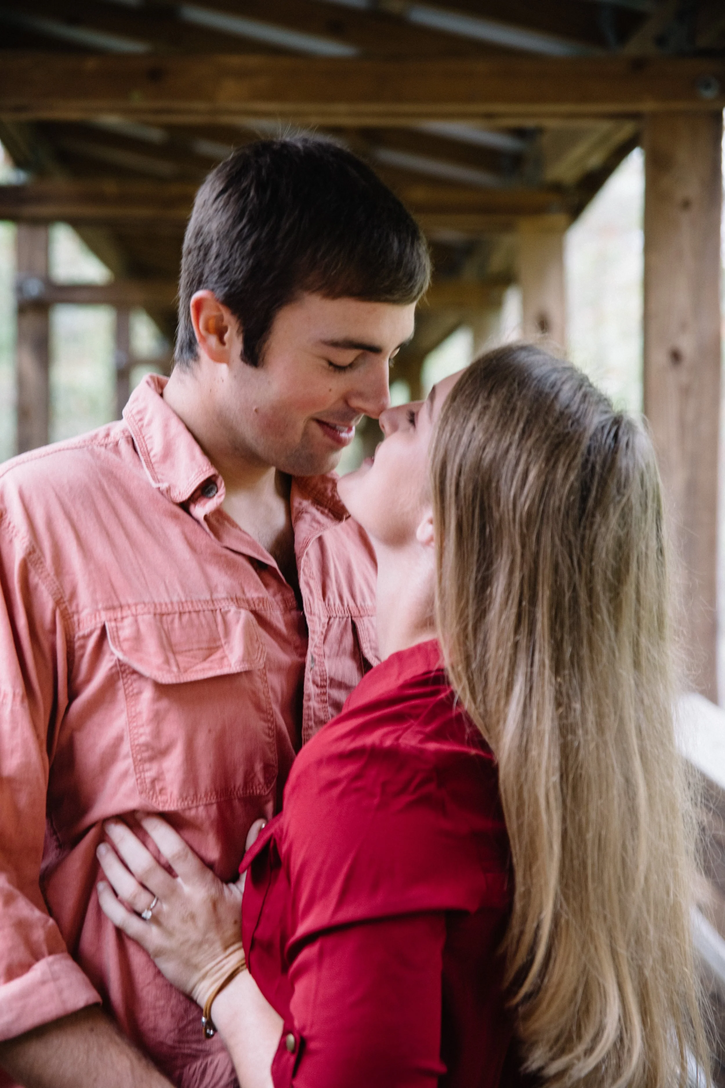 A couple is close together, smiling and looking into each other's eyes, with the man wearing a pink shirt and the woman wearing a red shirt, outdoors under a wooden structure.