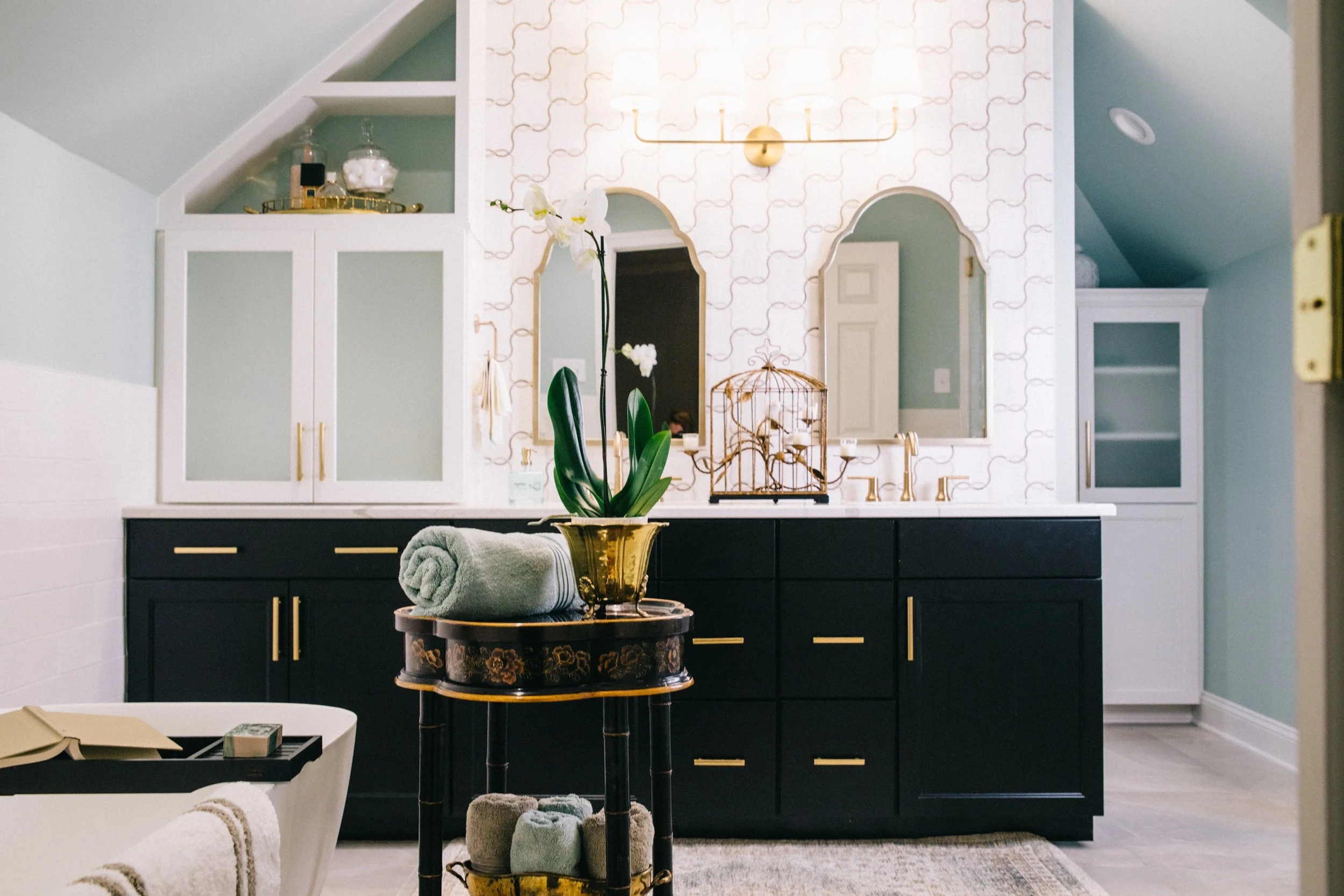 A decorated living room with a black sideboard, gold accents, a potted orchid, a birdcage, and a wall with light wallpaper and built-in shelves.