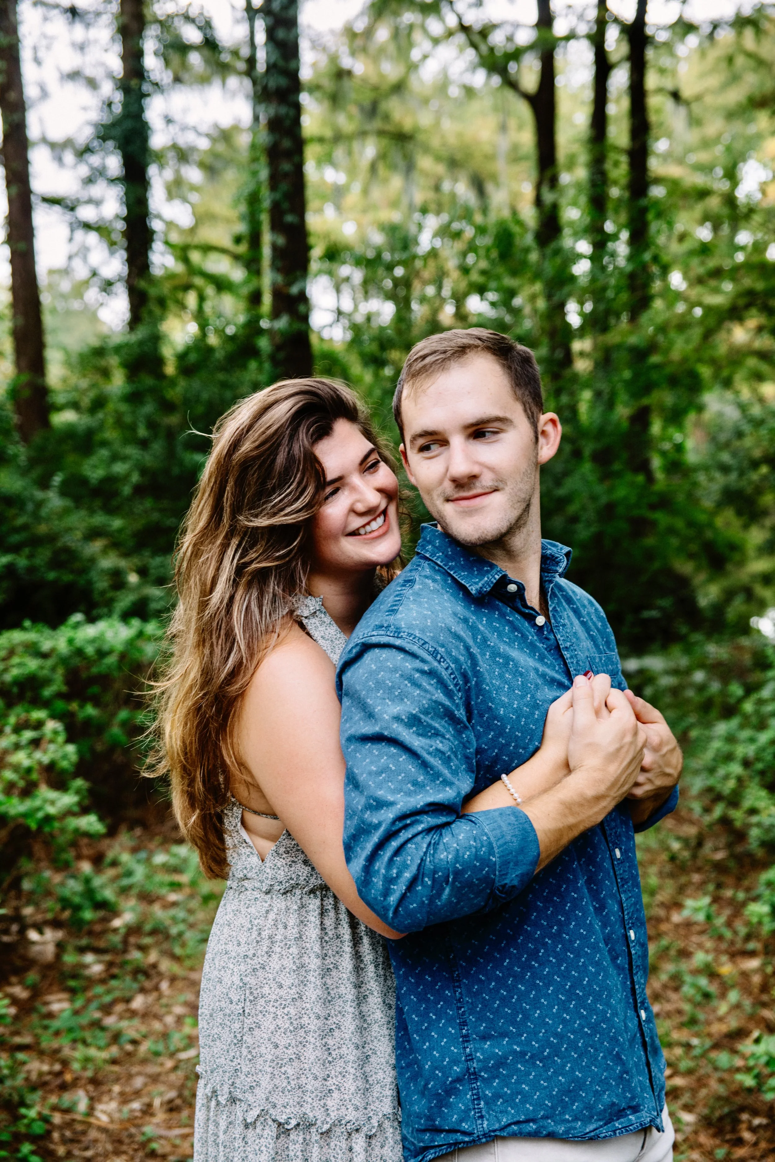 A couple standing close together in a forest, smiling and looking at each other, with the woman hugged from behind and the man looking to the side.