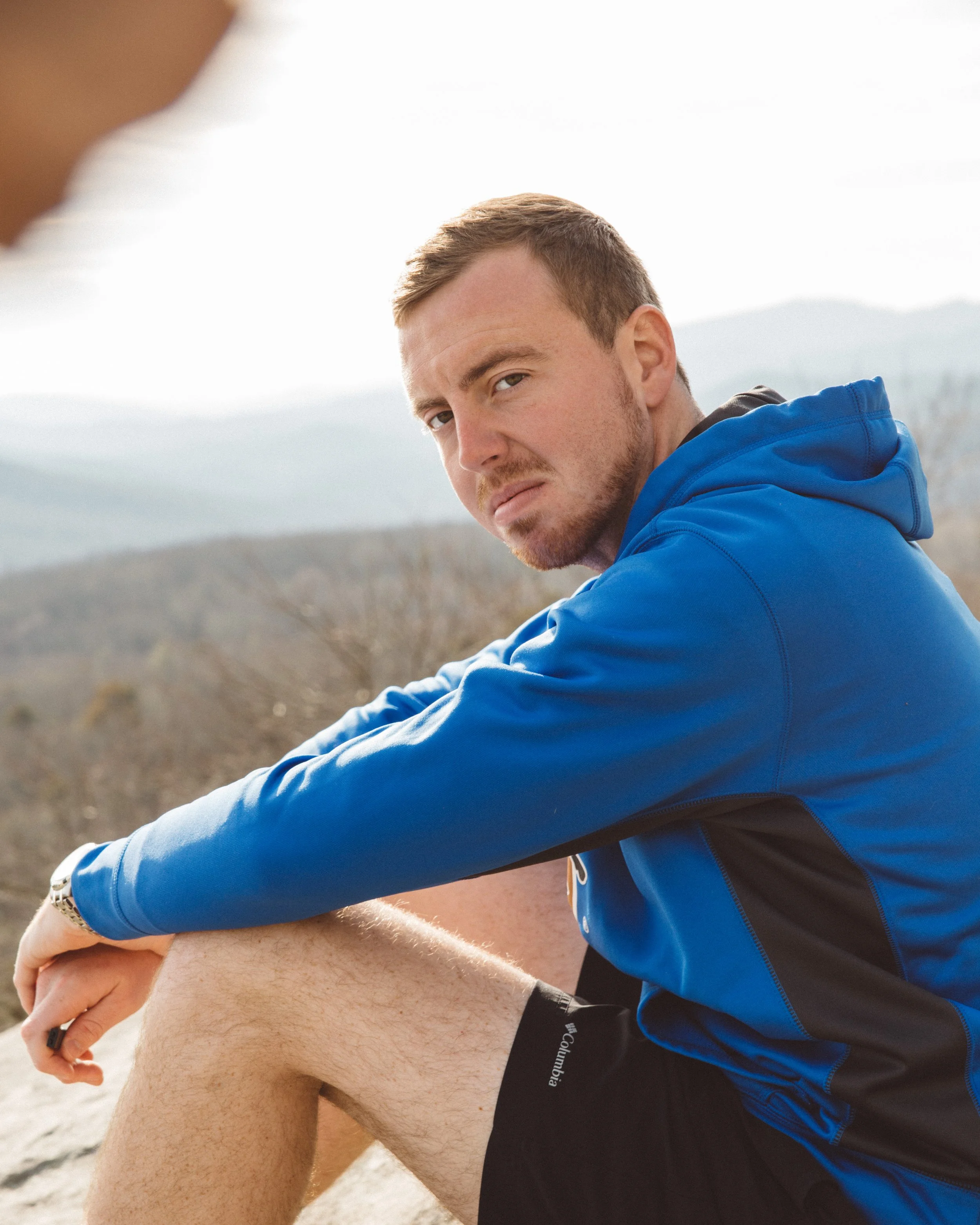 Man in blue jacket sitting outdoors, looking serious, with a mountain landscape in the background.